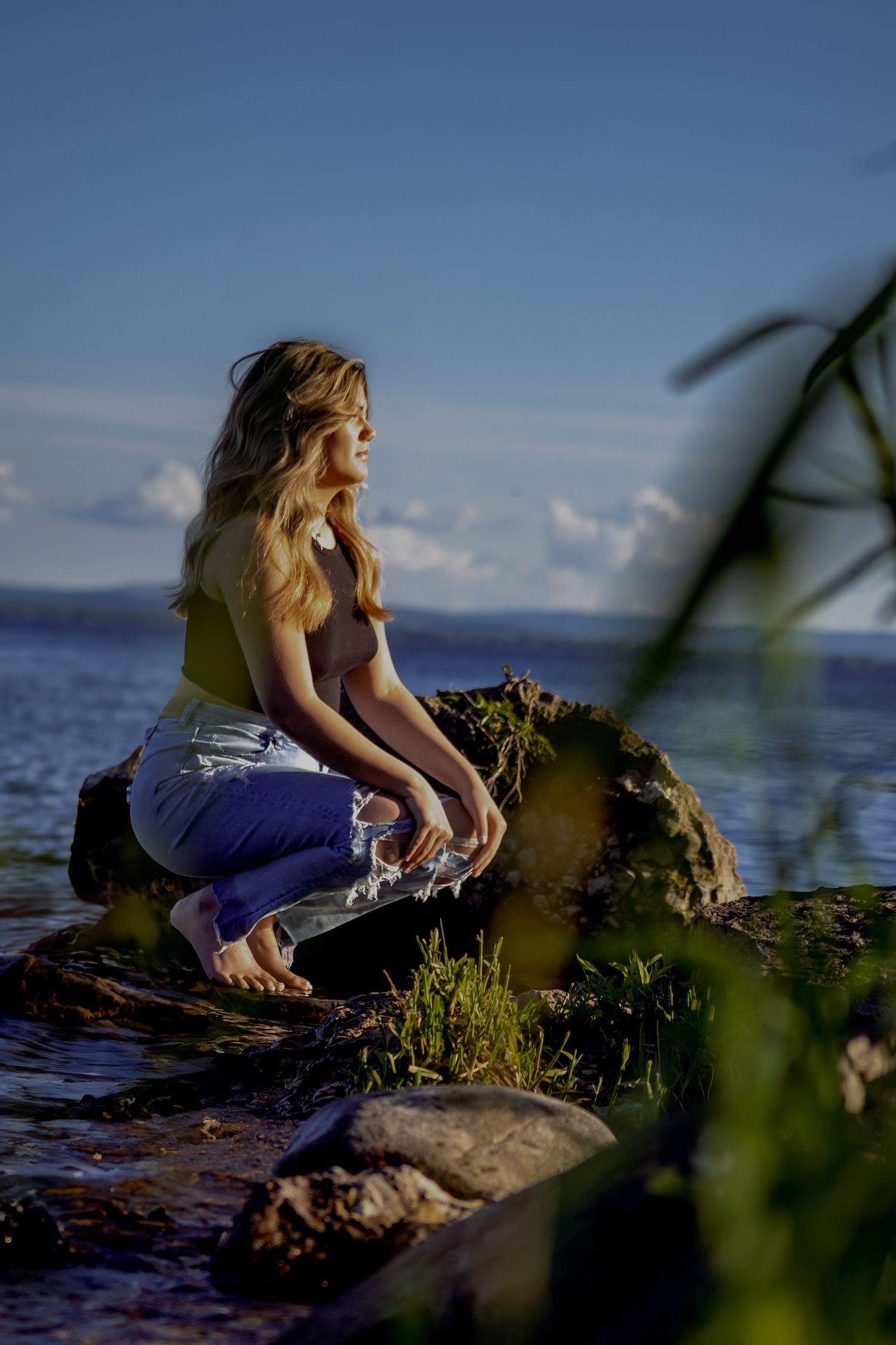 Young woman with wavy blonde hair sits barefoot on rocks by a serene lake, wearing jeans and a yellow top.