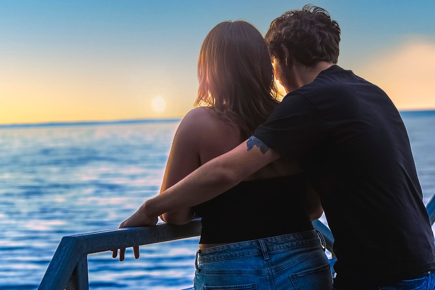 Couple embracing at sunset overlooking calm blue ocean waters, viewed from behind on a boat deck.