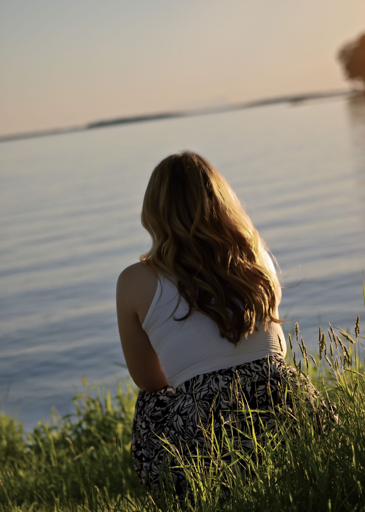 Young woman with long wavy hair sitting by a calm lake at sunset, wearing a white top and floral skirt.