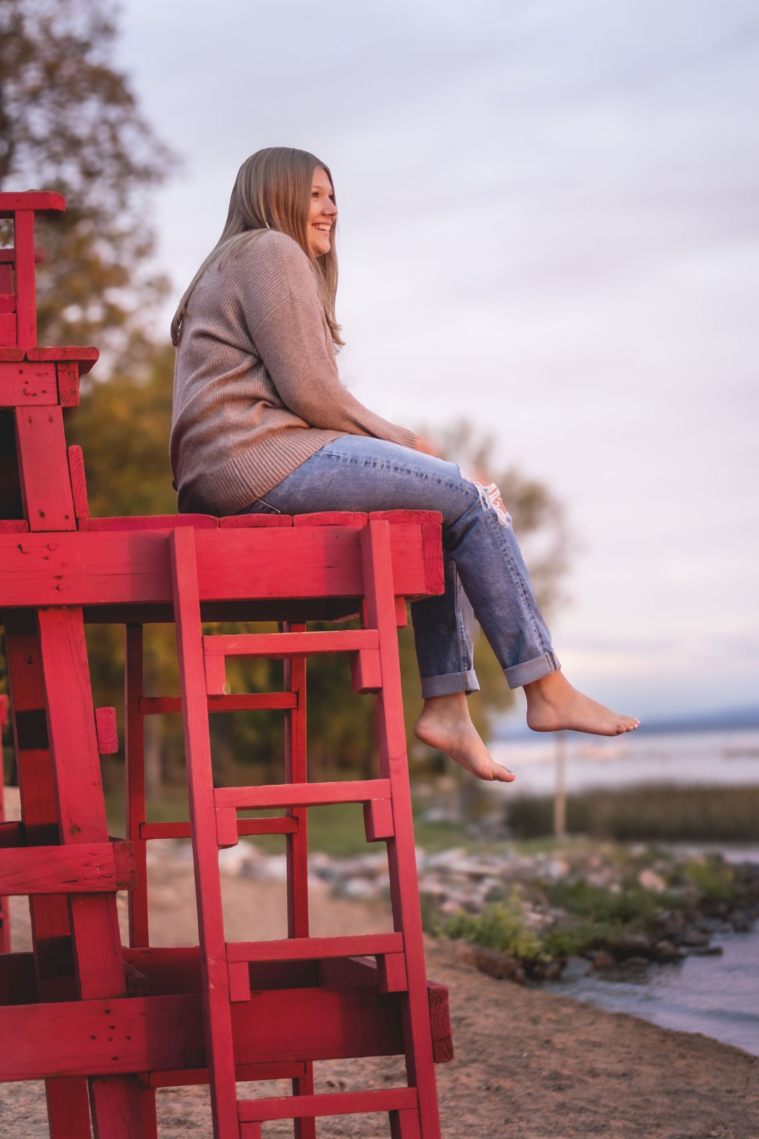 Smiling woman sitting barefoot on a red lifeguard stand near a beach at dusk, wearing jeans and a tan sweater.