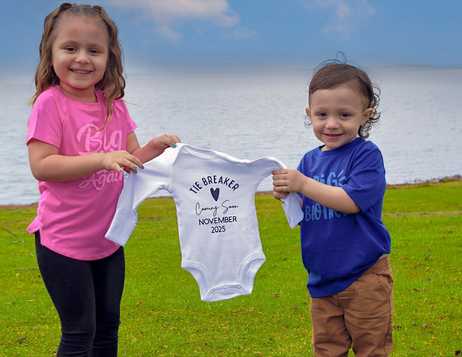 Two young children hold up a personalized baby announcement onesie outdoors near a bright blue waterfront.