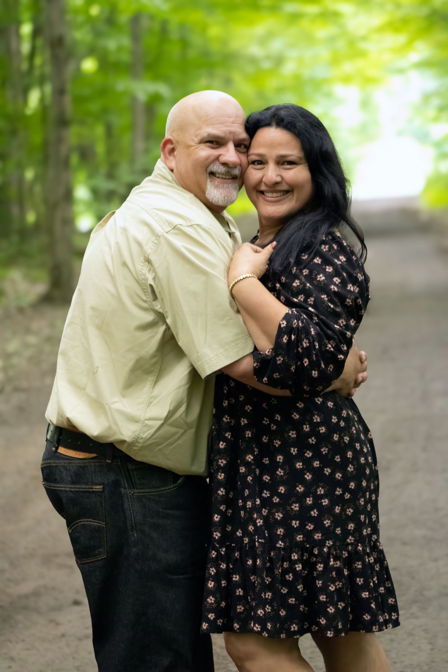 A smiling middle-aged couple embracing on a tree-lined path, woman in floral dress, man in light shirt.