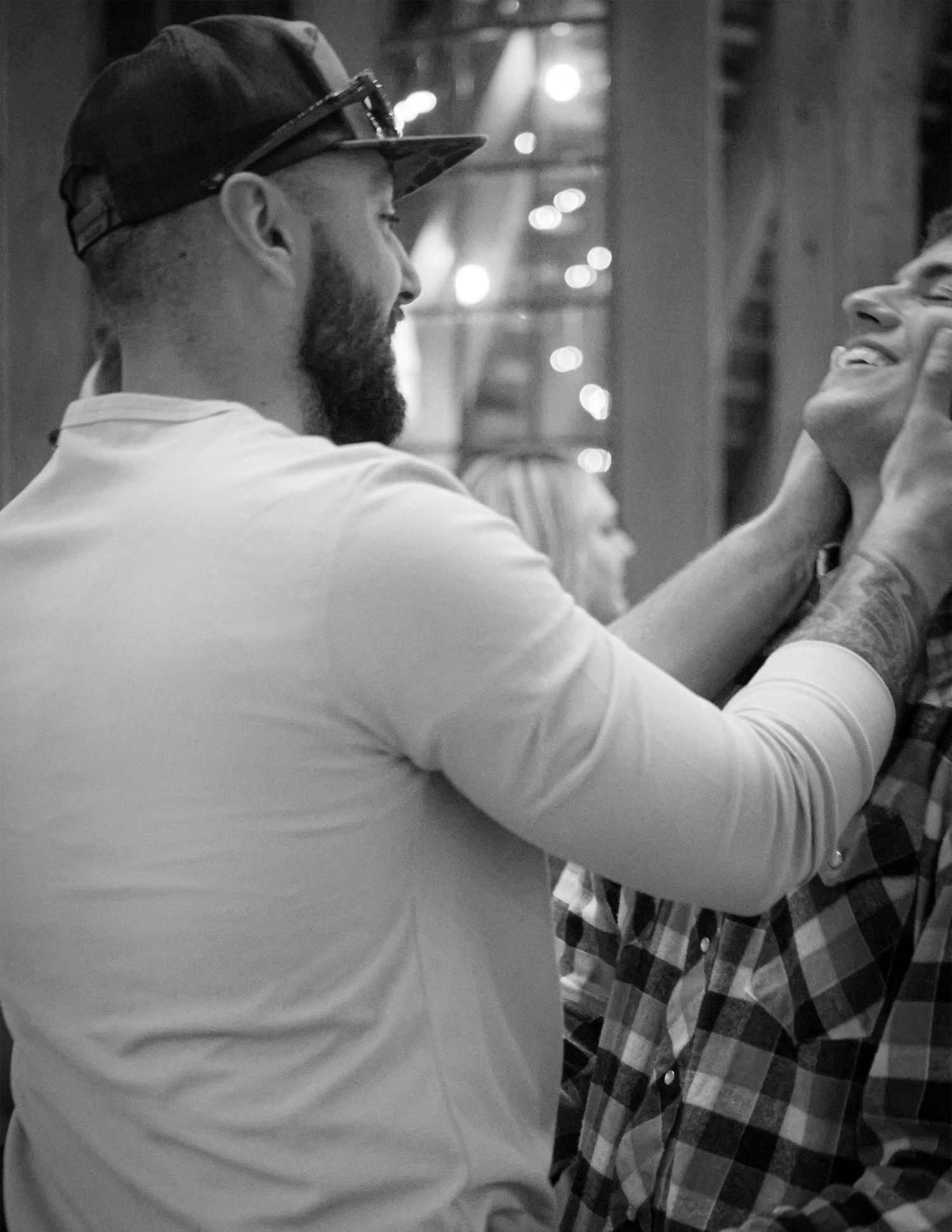 Black and white photo of two bearded men laughing together, one wearing a cap and white shirt, the other in a plaid shirt.