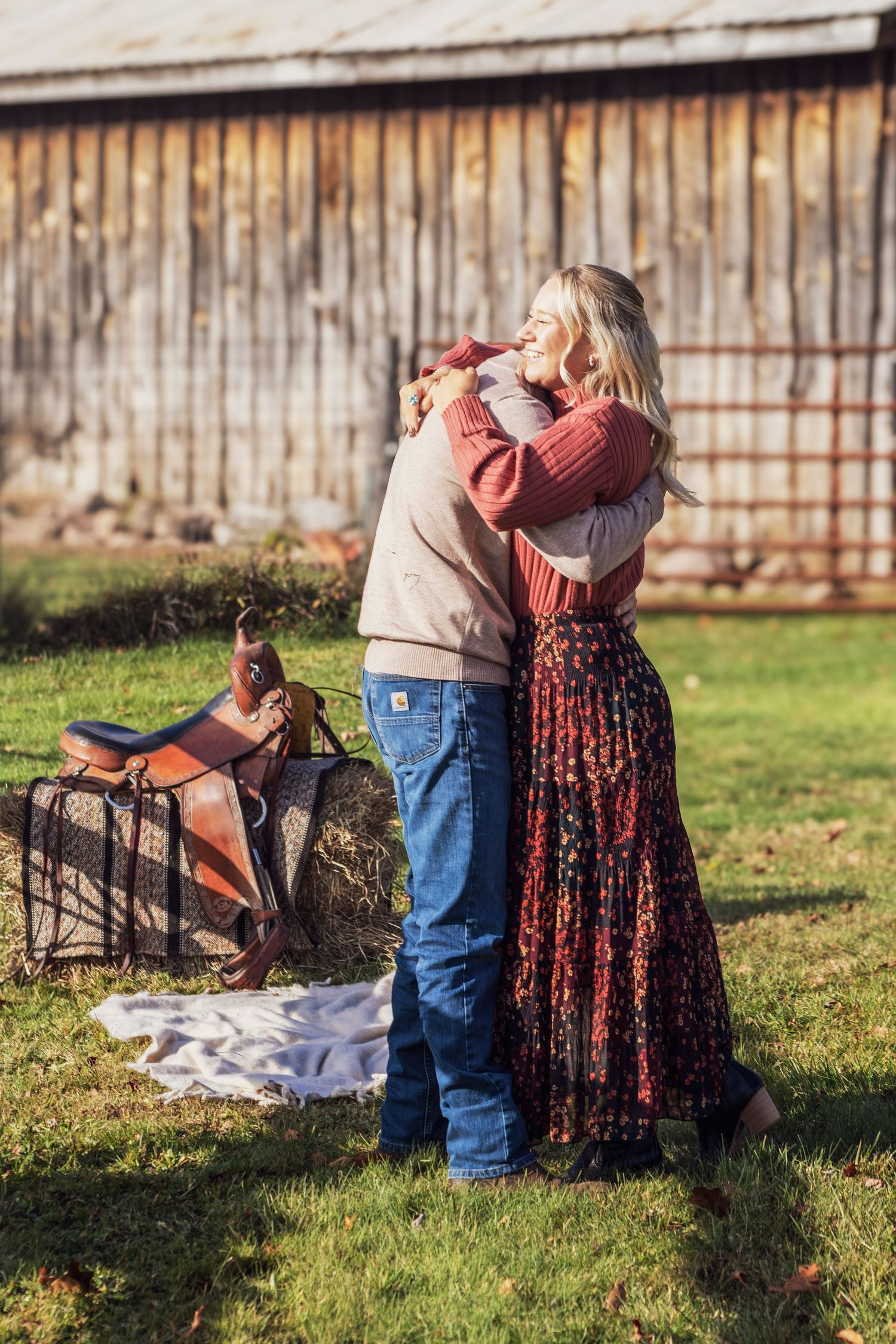 Couple embracing in a rustic farm setting with a horse saddle and hay bale, wearing casual country attire.