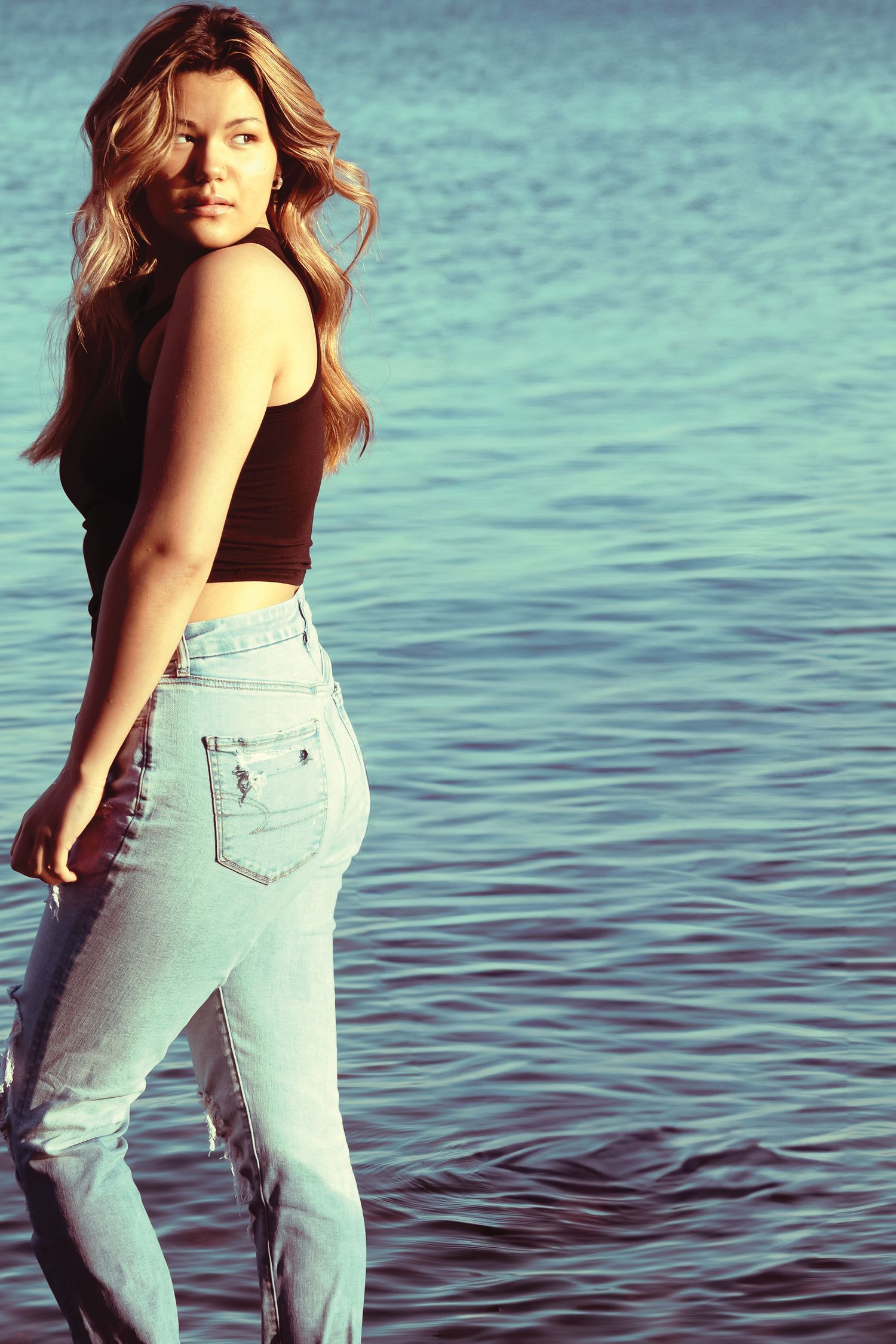 Young woman with long hair in black crop top and light blue jeans posing by calm blue water on a sunny day.