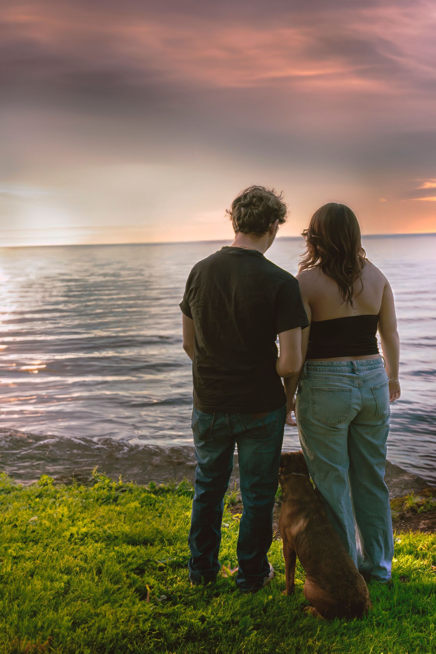Couple and dog watching a dramatic sunset over the ocean from a grassy cliff edge.