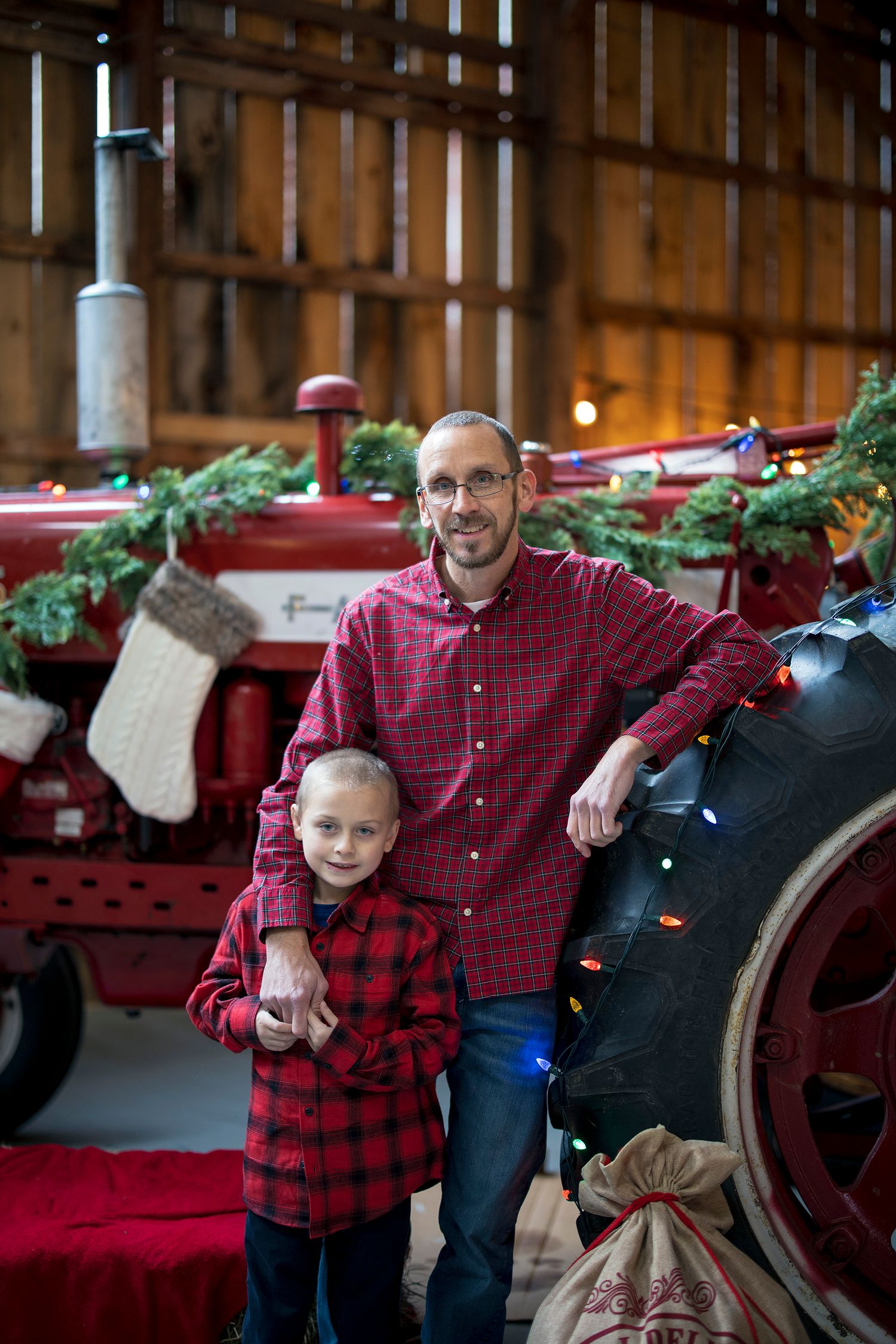 Father and son in matching red plaid shirts pose by a decorated vintage red tractor in a rustic barn at Christmas.