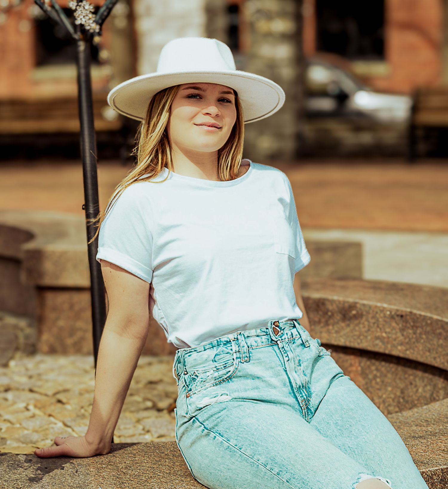 Young woman in white hat, white t-shirt, and light jeans sitting outdoors in a sunny urban setting.