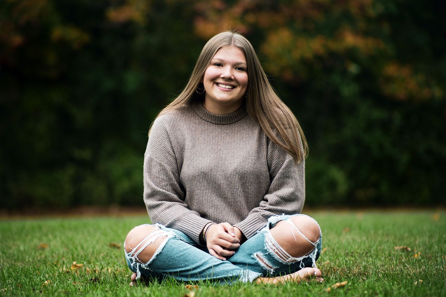 Smiling young woman sitting cross-legged on grass in ripped jeans and gray sweater with colorful fall trees behind her.