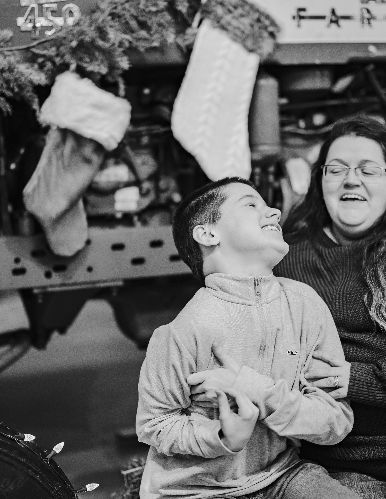 Laughing boy and woman sit together near Christmas decorations with hanging stockings in a black and white holiday photo.
