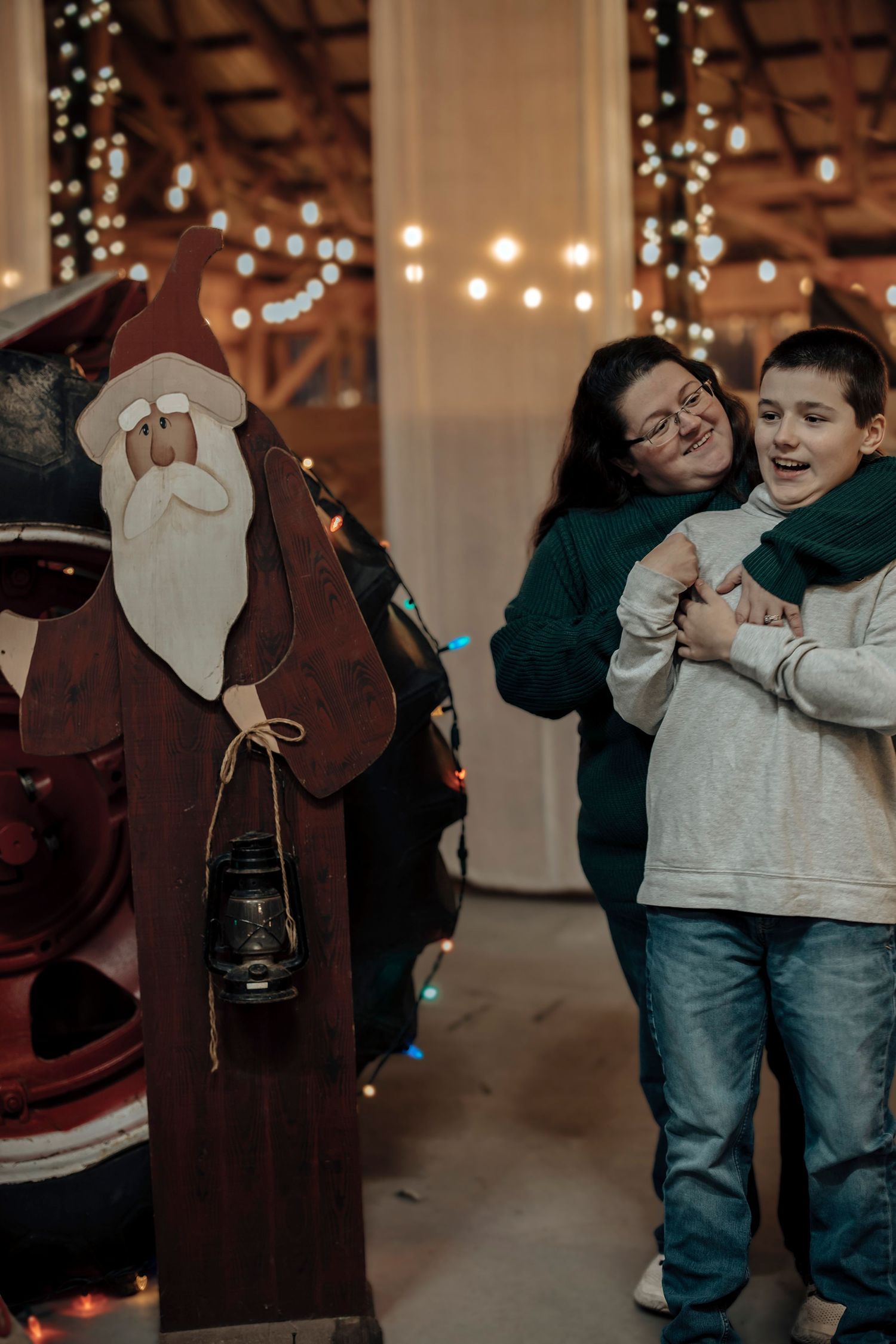 Mother and son share a joyful hug beside a wooden Santa Claus decoration with holiday lights in background.