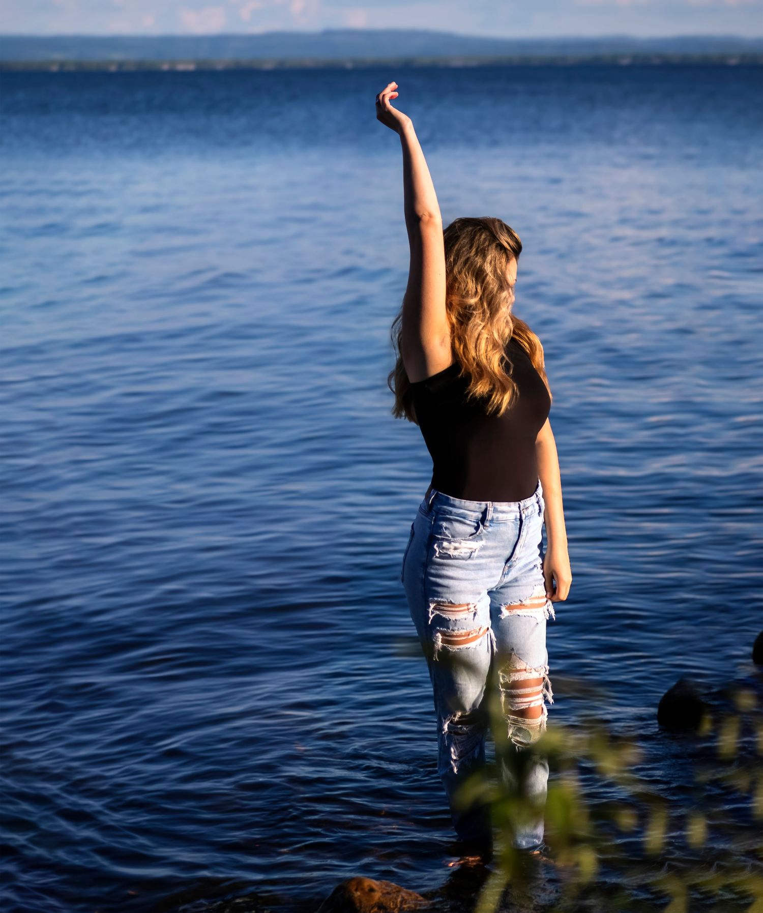Young woman in black top and ripped jeans stands in shallow lake water with one arm raised triumphantly.