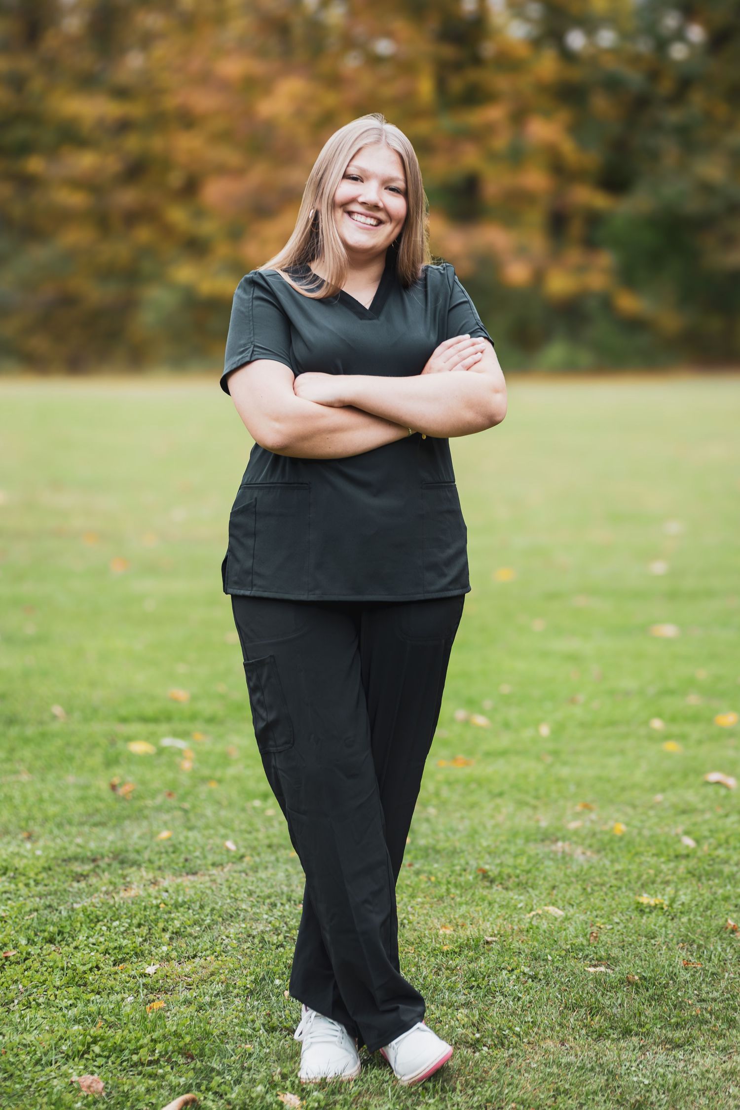 Smiling blonde woman in black scrubs stands outdoors with arms crossed on green grass with autumn trees behind her.
