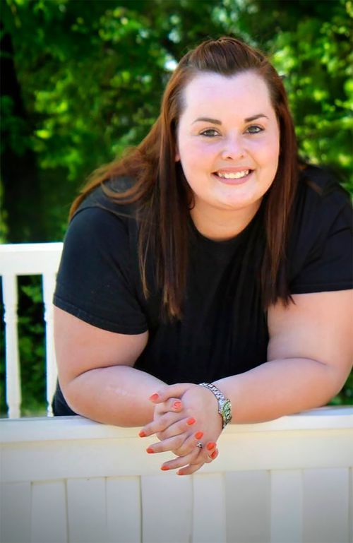 Smiling young woman with auburn hair leaning on white railing outdoors, wearing black t-shirt with green trees behind her.