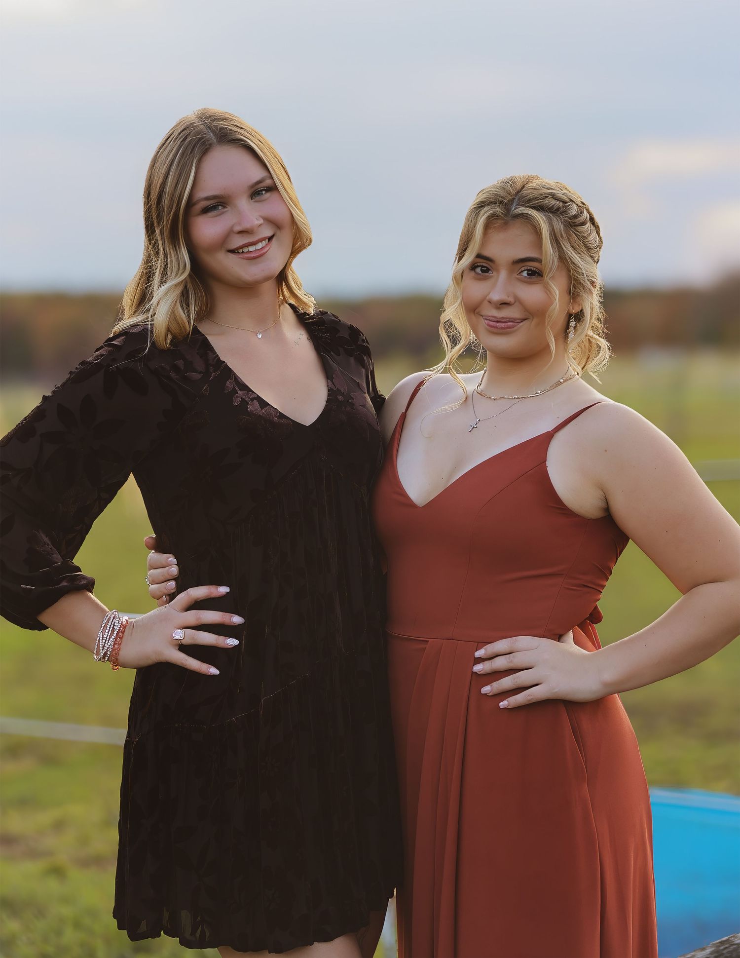 Two young women pose outdoors in elegant dresses, one black and one rust-colored, against an open field.