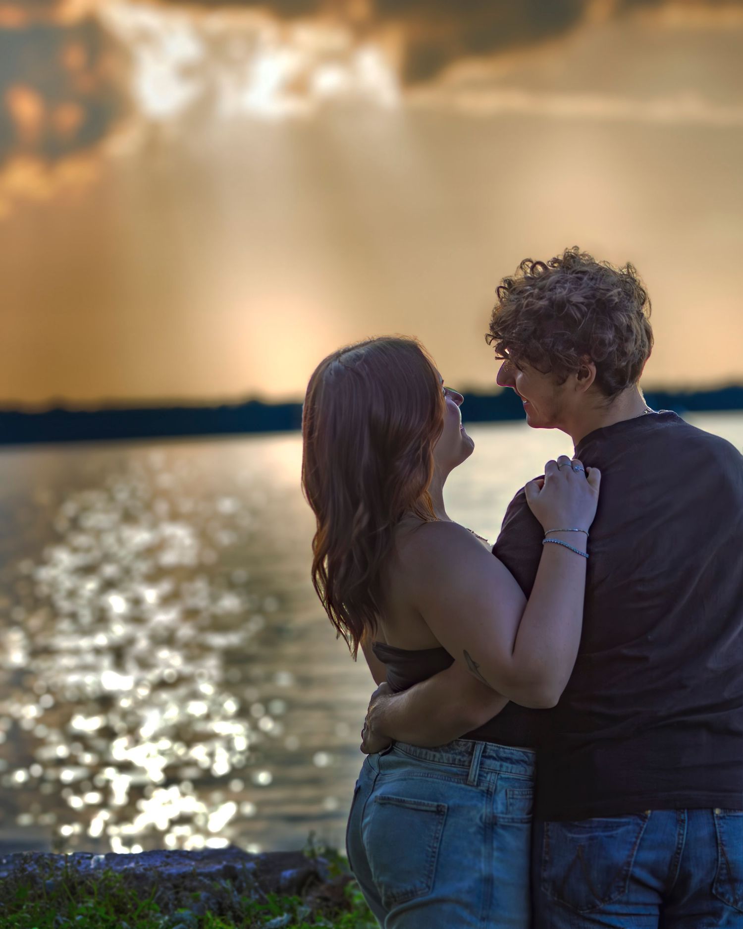 Couple embracing by a shimmering lake at sunset with dramatic golden rays breaking through storm clouds.