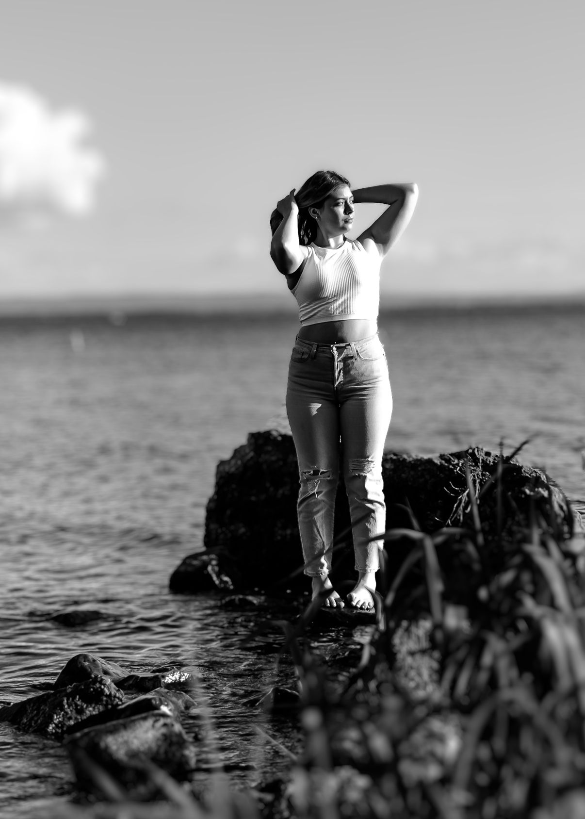 Black and white photo of a woman posing on rocks by the ocean, hands behind her head, wearing jeans and a white top.