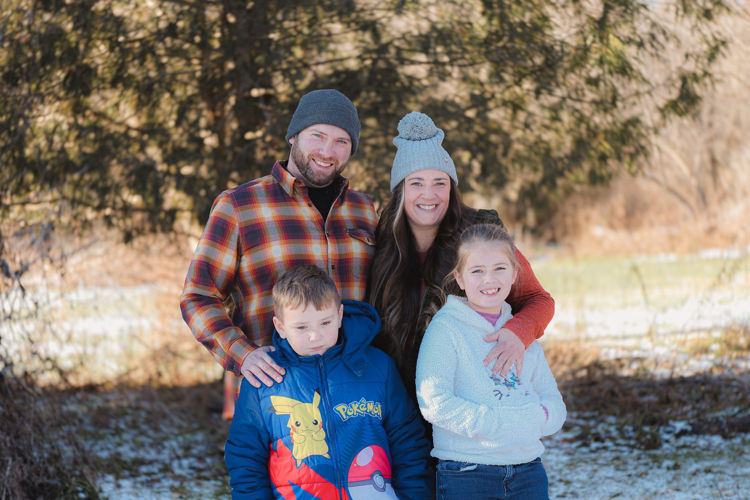 Family of four wearing winter clothing and hats posing together outdoors in a snowy wooded area.