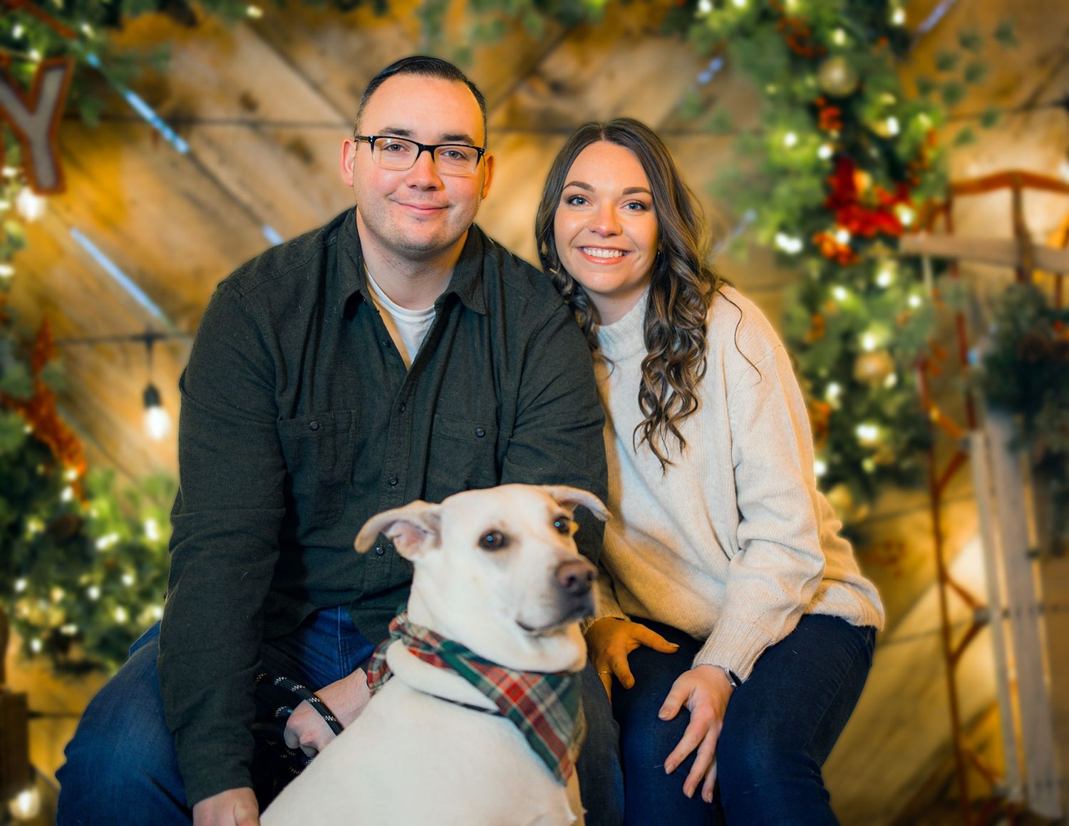 Smiling couple poses with their white dog in front of a festive Christmas tree holiday portrait backdrop.