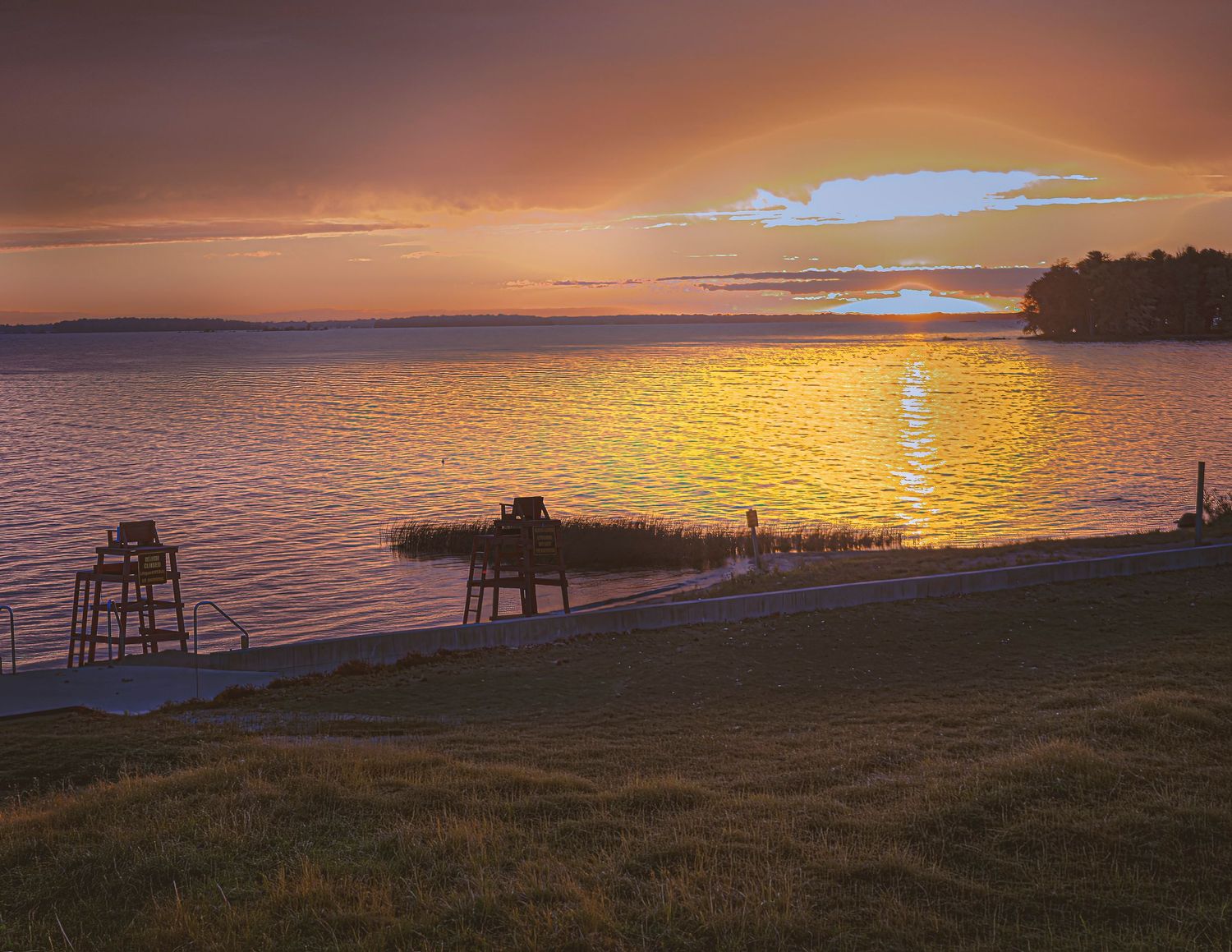 Golden sunset over a calm lake with a lifeguard chair and dock silhouetted against the glowing sky.