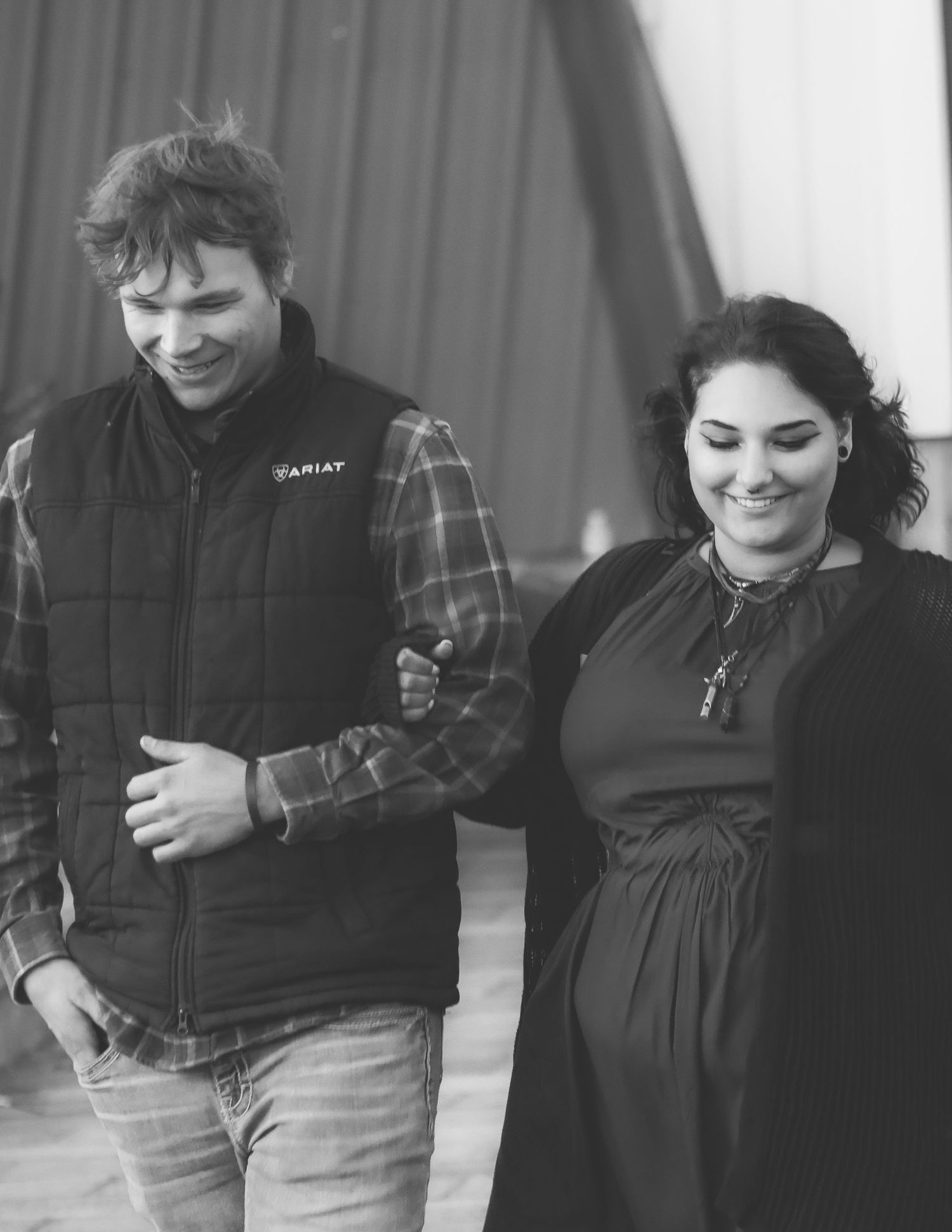 Black and white photo of a smiling pregnant woman walking arm-in-arm with a man wearing an Ariat vest.