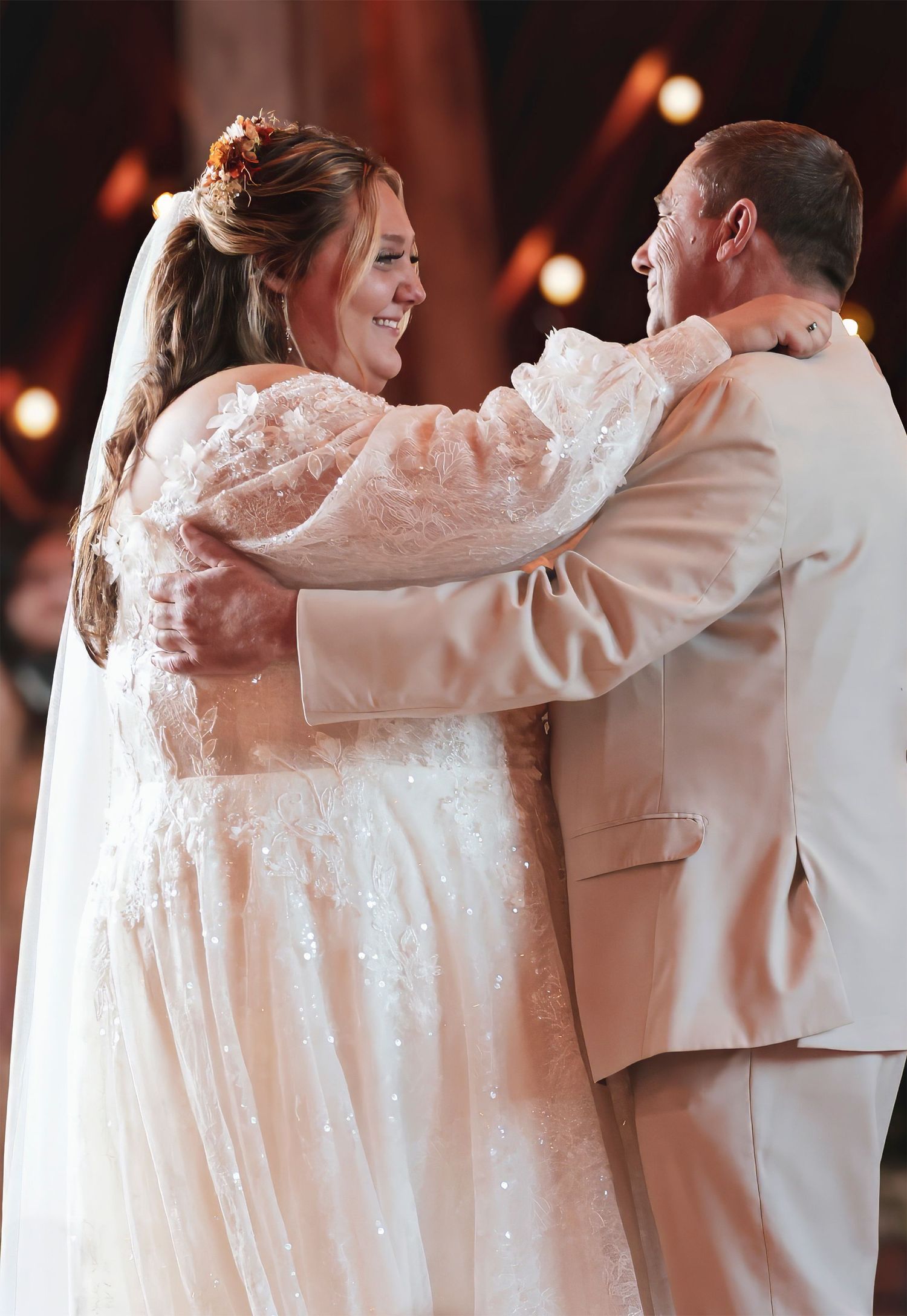 Bride and groom share first dance, smiling joyfully, bride in lace gown with veil, groom in tan suit.