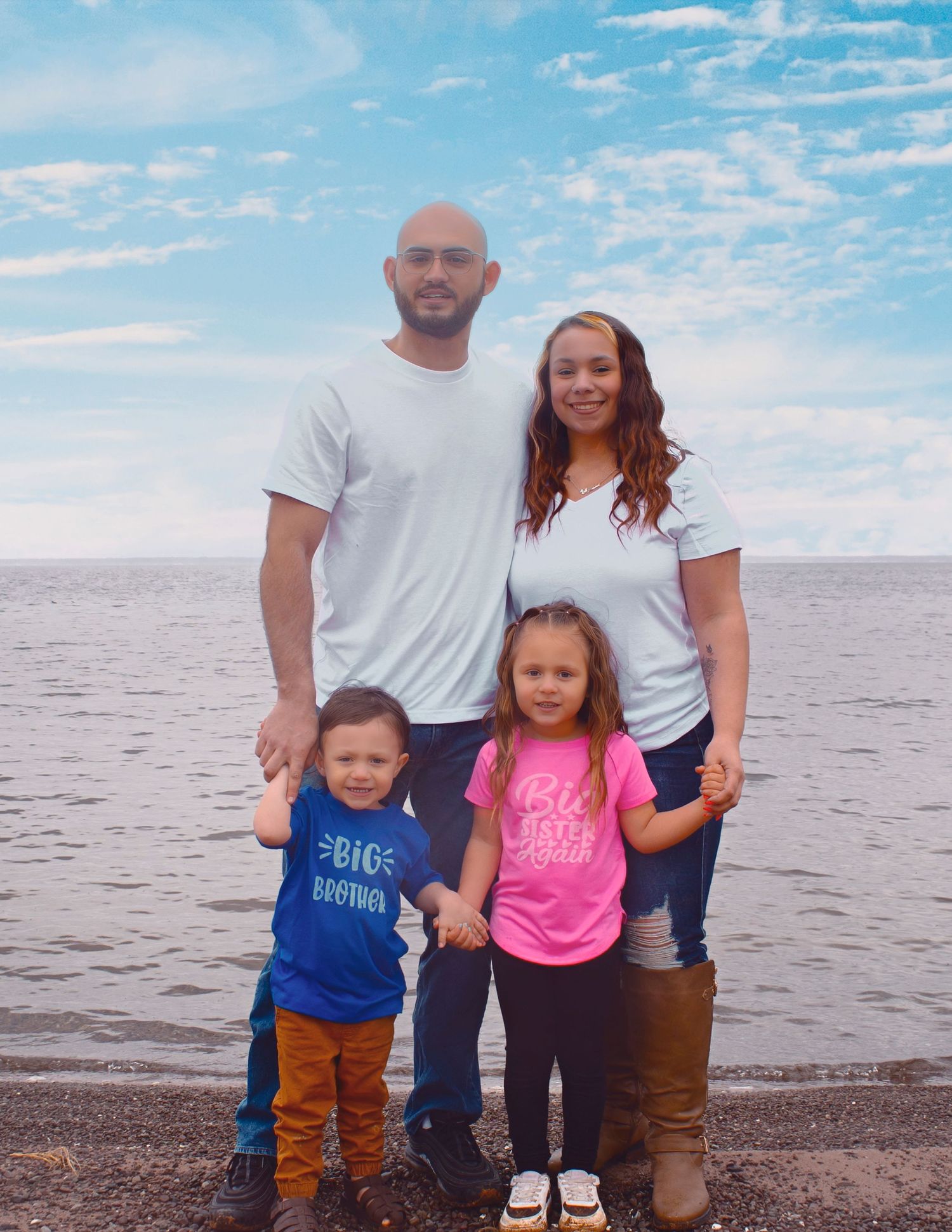 Family of four posing together on a rocky beach shoreline with blue sky and calm water in the background.