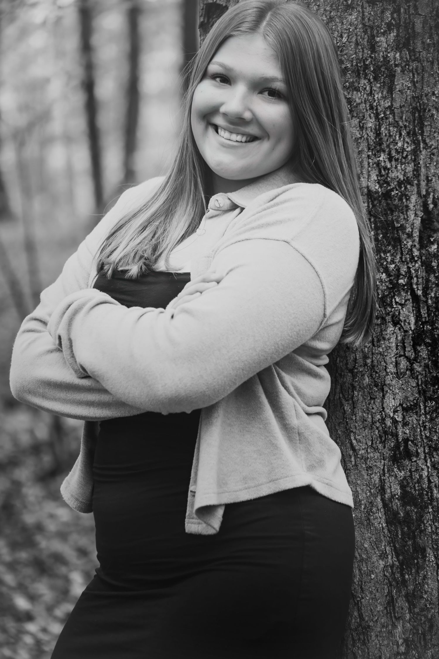 Smiling teenage girl with long hair leans against a tree with arms crossed in black and white forest photo.
