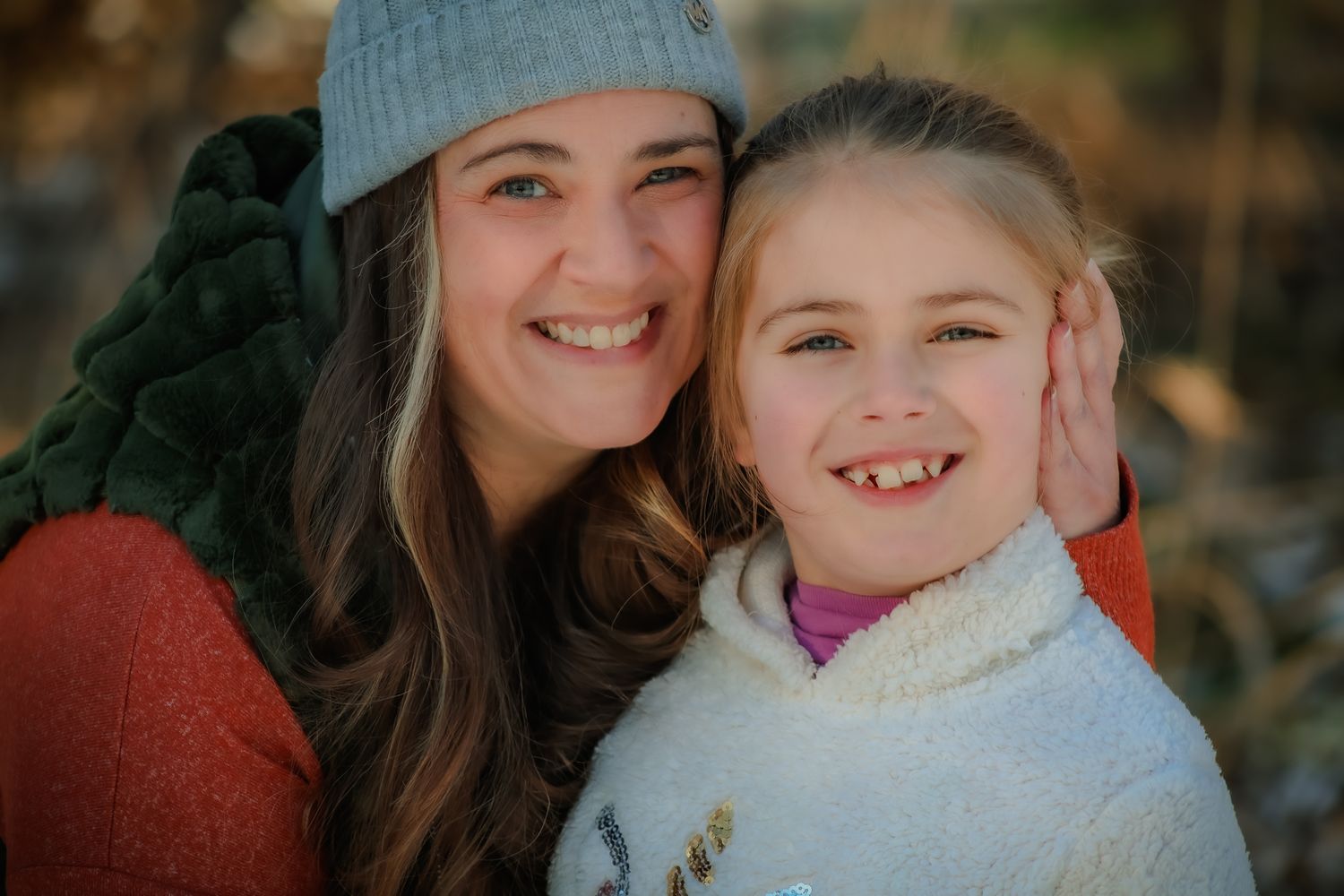 Smiling mother and young daughter posing cheek to cheek outdoors in warm winter clothing.