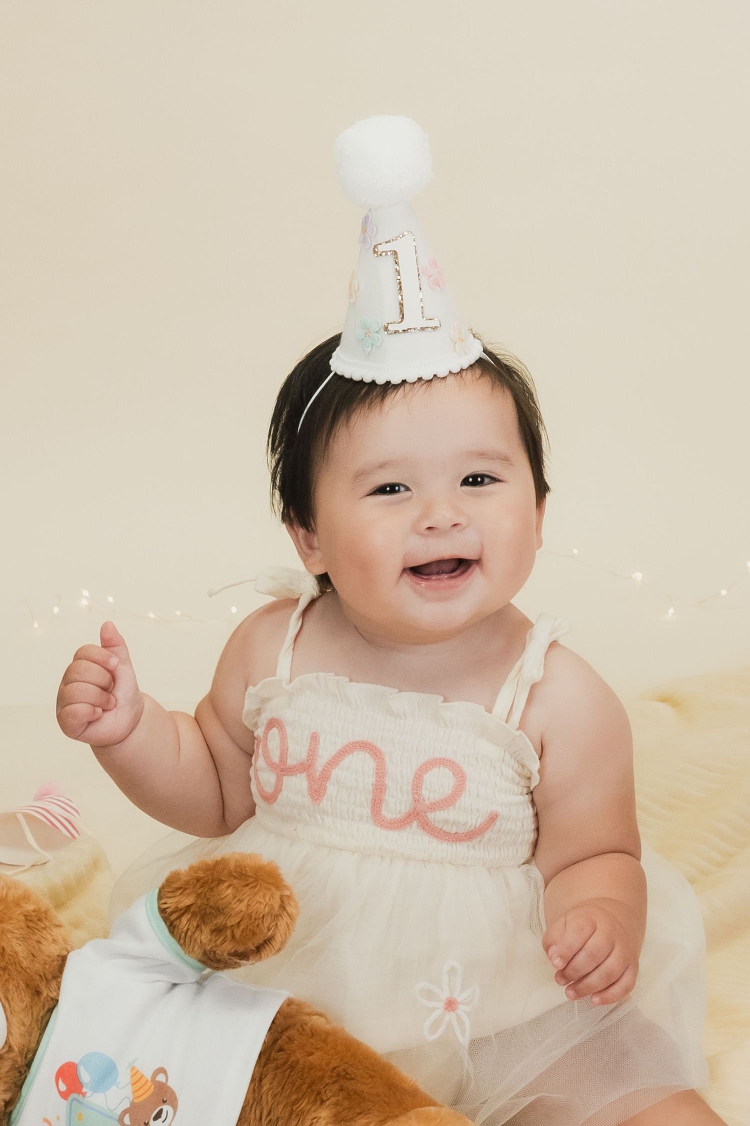 Smiling baby girl in white tutu dress and number one birthday hat celebrates her first birthday with teddy bear.