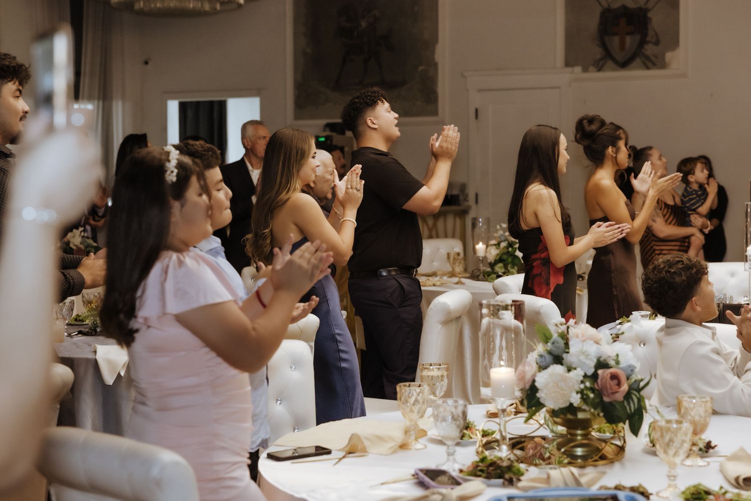 Wedding reception guests standing and applauding in an elegant venue with floral centerpieces.