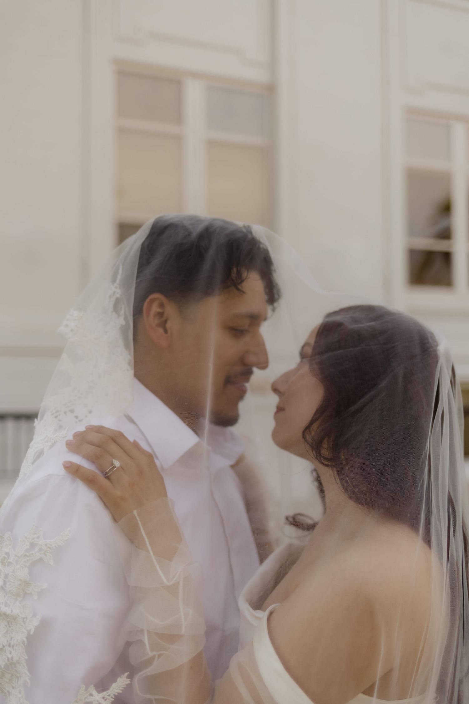 Romantic wedding portrait of bride and groom nearly kissing under a flowing lace veil outside a white building.