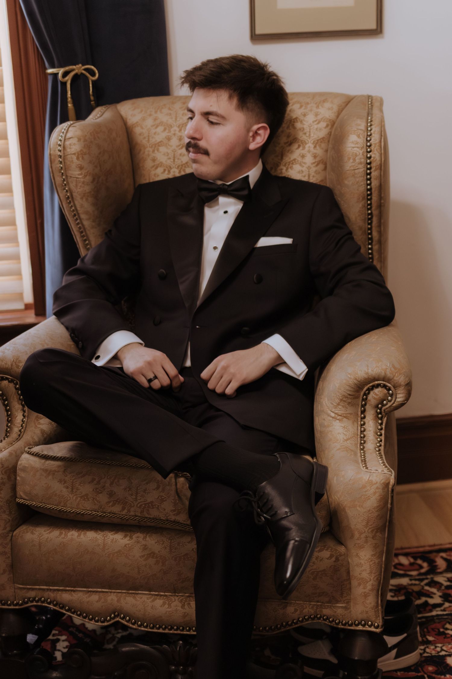 Groom in black tuxedo with bow tie relaxing in a vintage gold armchair in an elegant room.