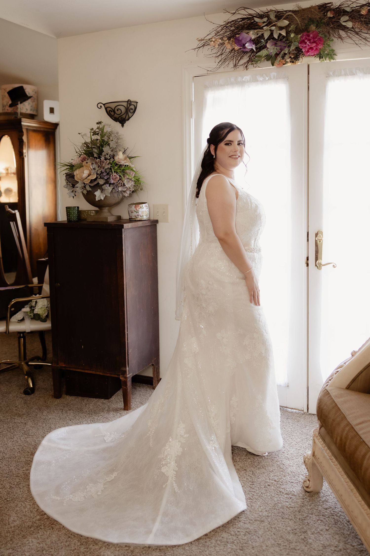 Bride in a lace mermaid wedding gown with long train stands elegantly by a bright window before her ceremony.