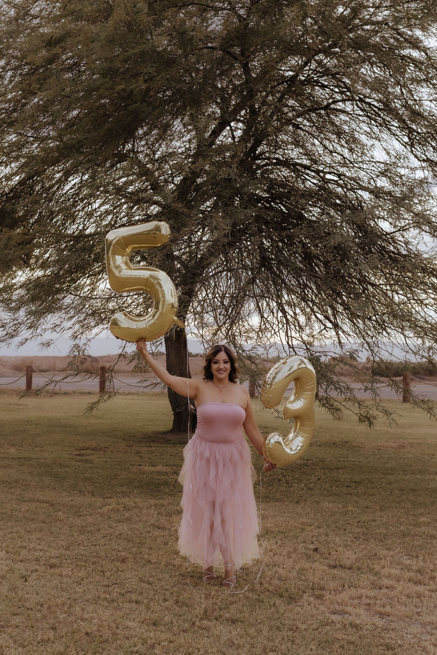 Woman in pink tulle dress holding gold number 53 balloons outdoors near a tree.