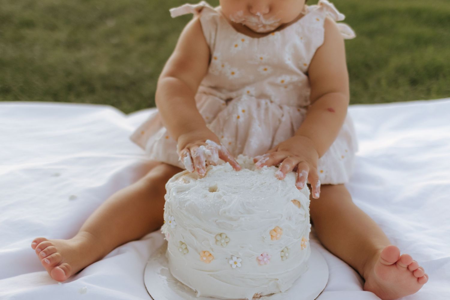 Baby sitting on white blanket smashing hands into white birthday cake outdoors.