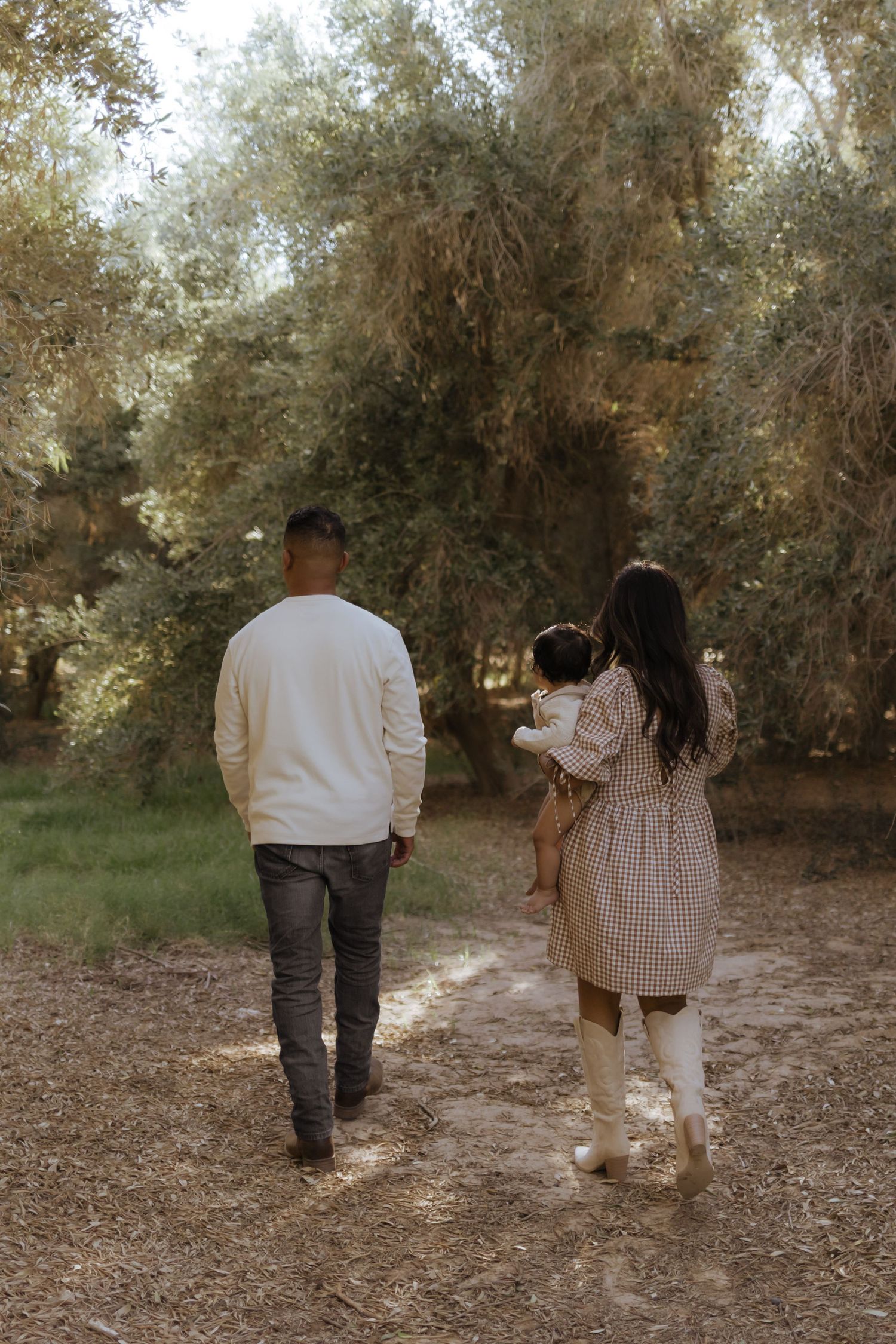 Young family walking through sunlit grove of trees on dirt path from behind.