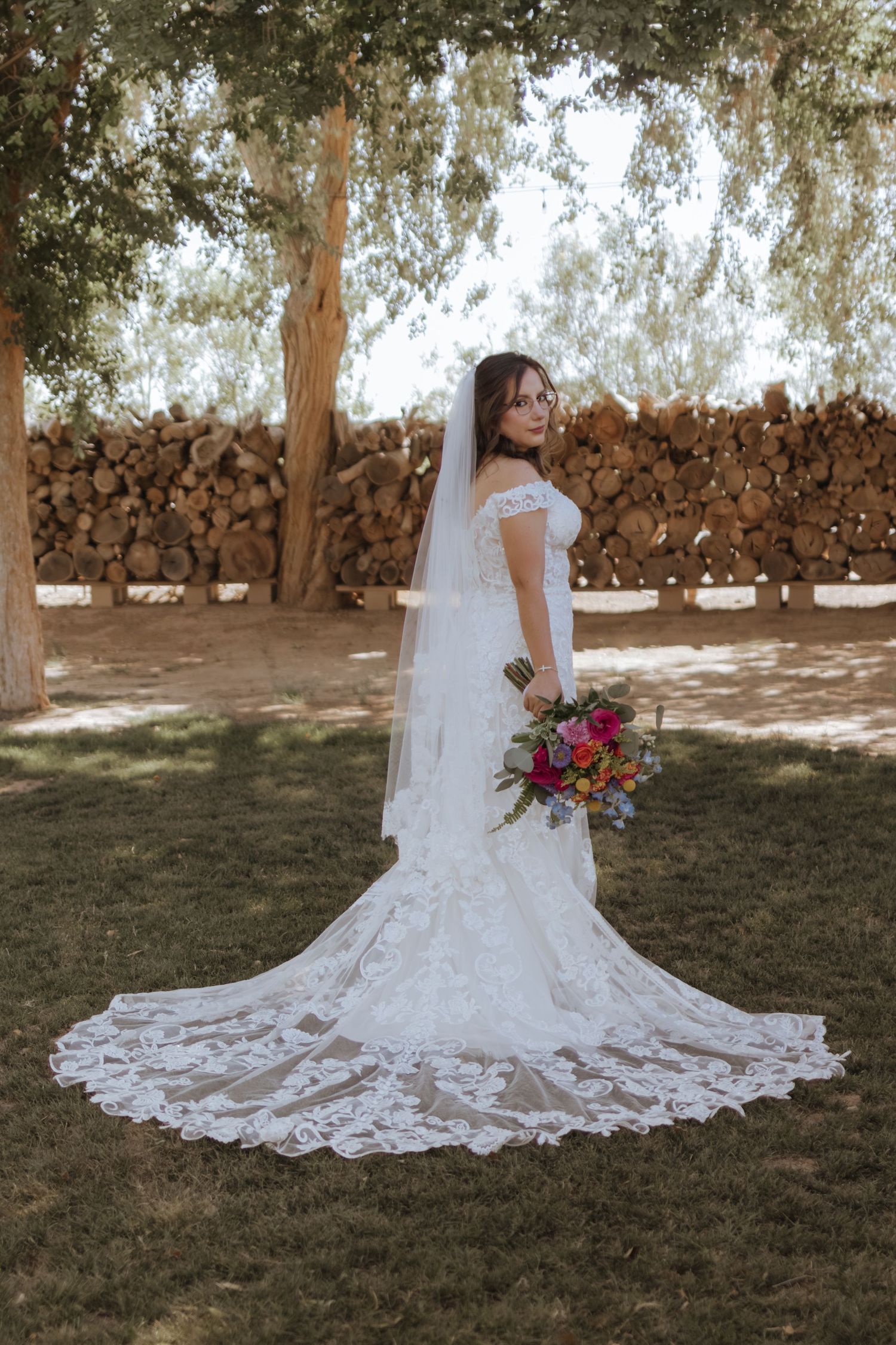 Bride in off-shoulder lace gown with cathedral veil holds colorful bouquet while posing near rustic stacked wood logs.