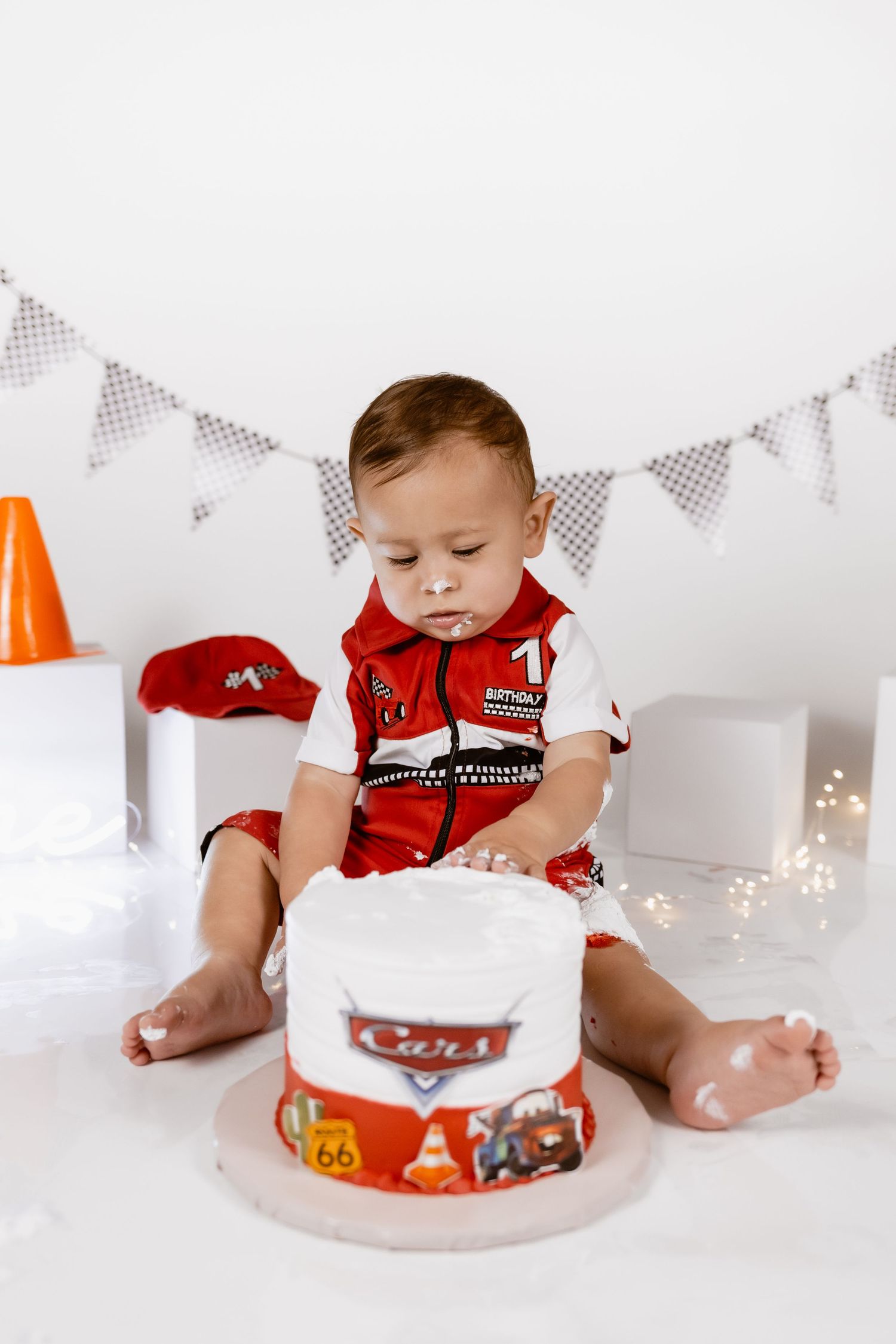 Baby boy in racing outfit celebrating first birthday with a Cars themed smash cake on white background.