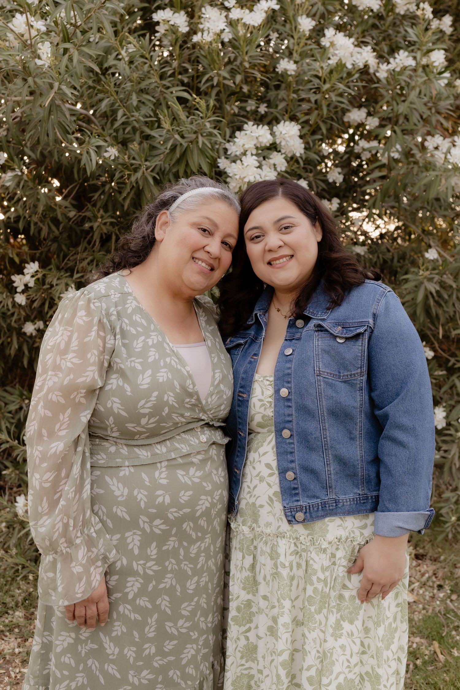 Two smiling women pose together in front of a lush flowering bush in soft natural light.