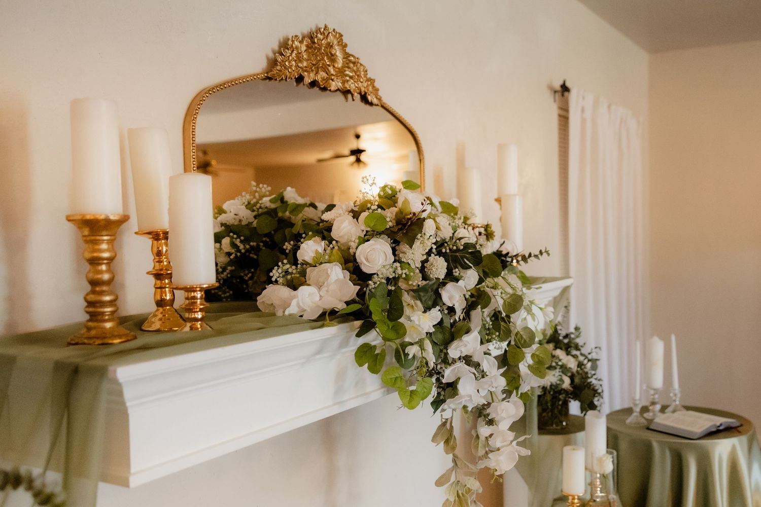 White floral arrangement with greenery adorns a white fireplace mantel with gold candlesticks in a wedding venue.