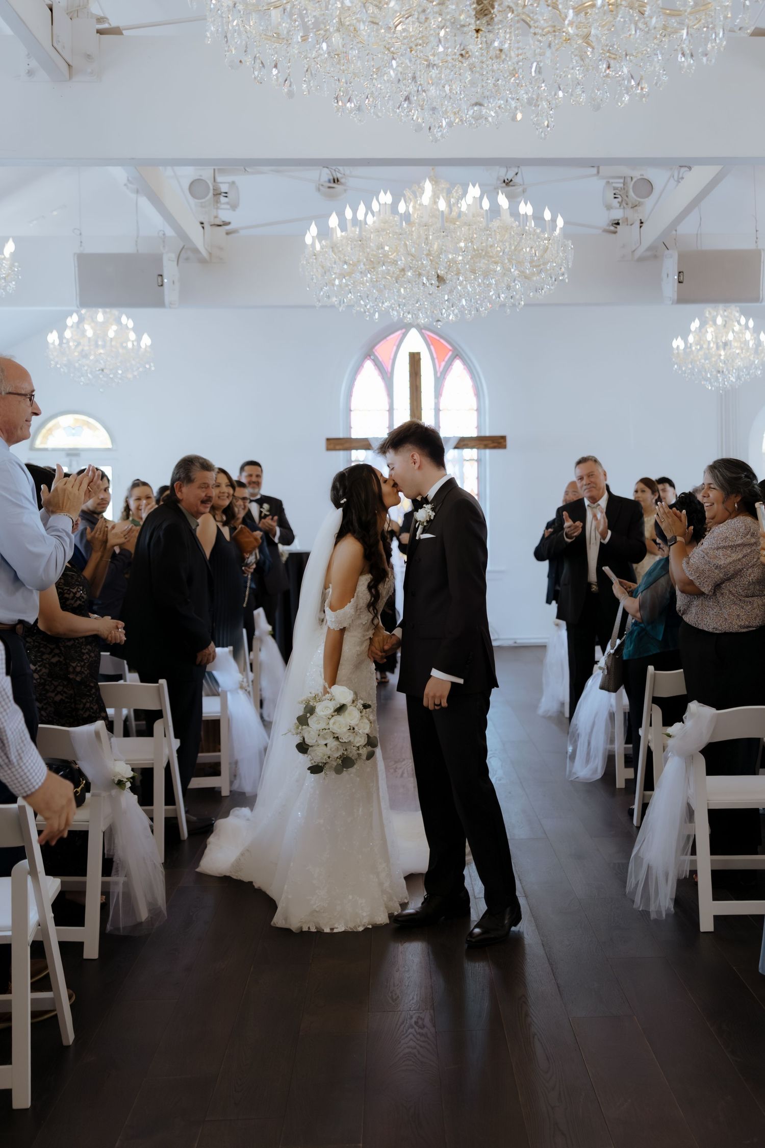 Bride and groom share first kiss walking down aisle in elegant white church venue with crystal chandeliers.