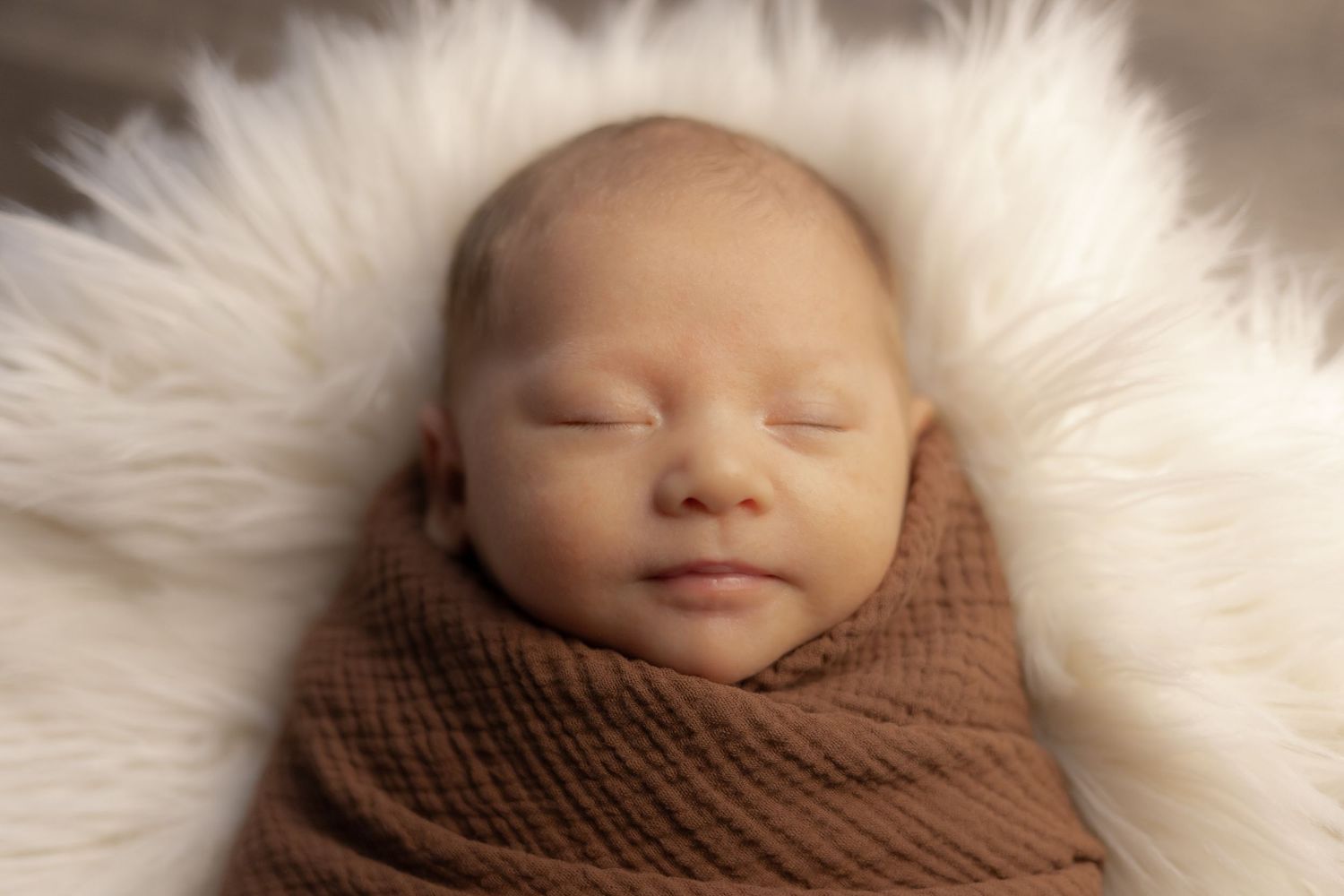 Peaceful newborn baby swaddled in brown wrap, sleeping on fluffy white fur blanket in professional photo.