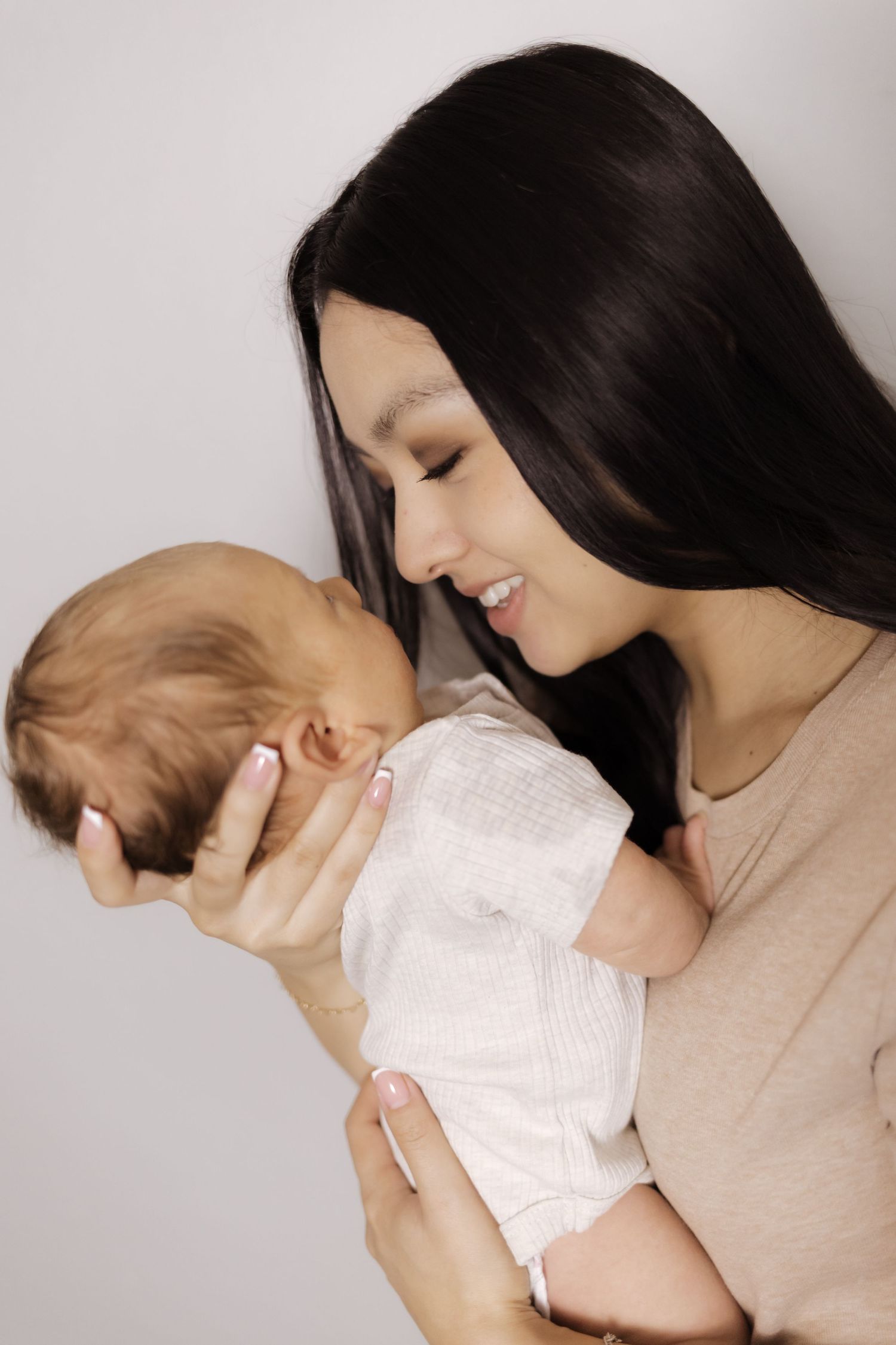 A smiling dark-haired mother tenderly holds her newborn baby close, gazing lovingly at the infant.