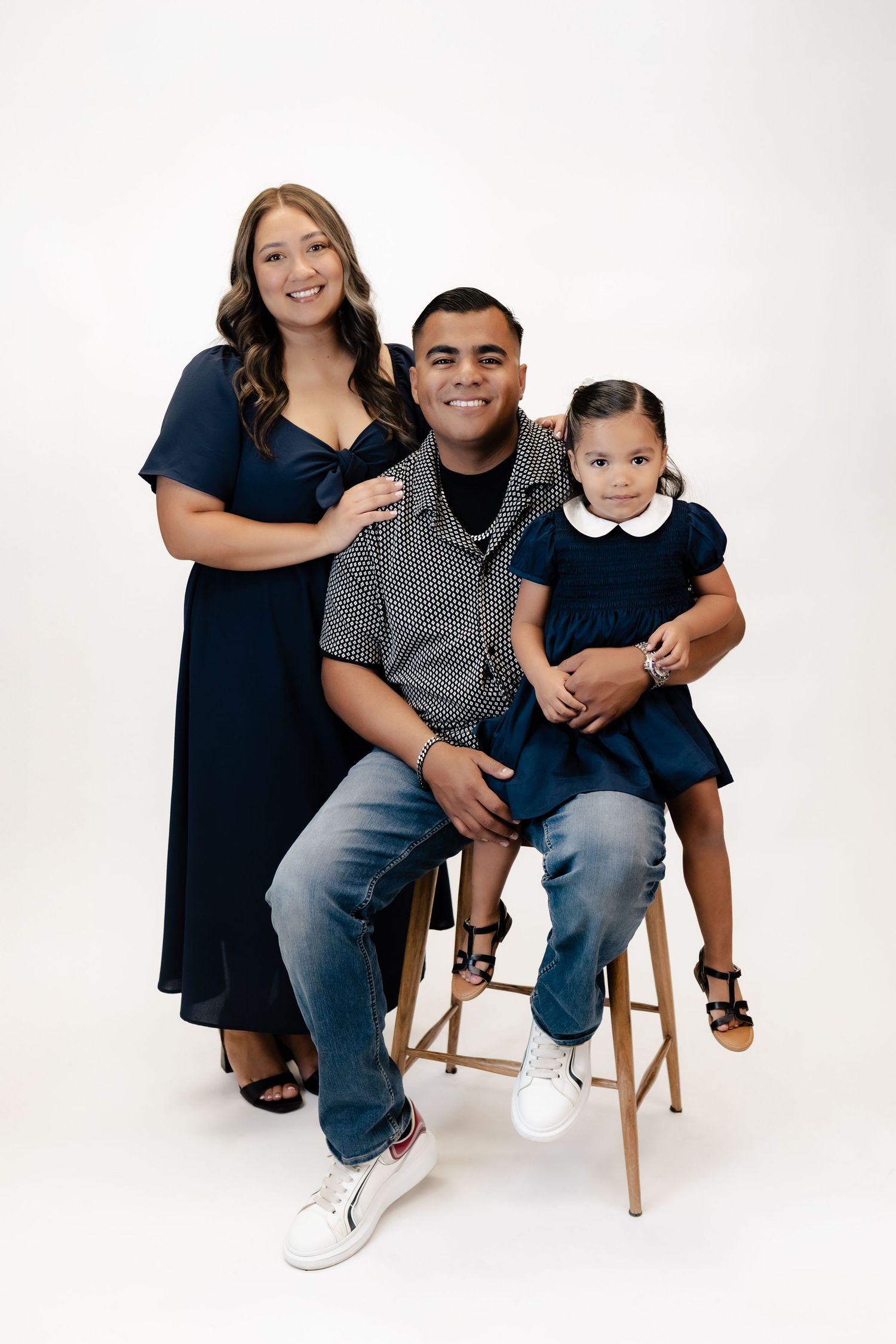 Family portrait of parents in navy outfits with toddler daughter on dad's lap against white background.
