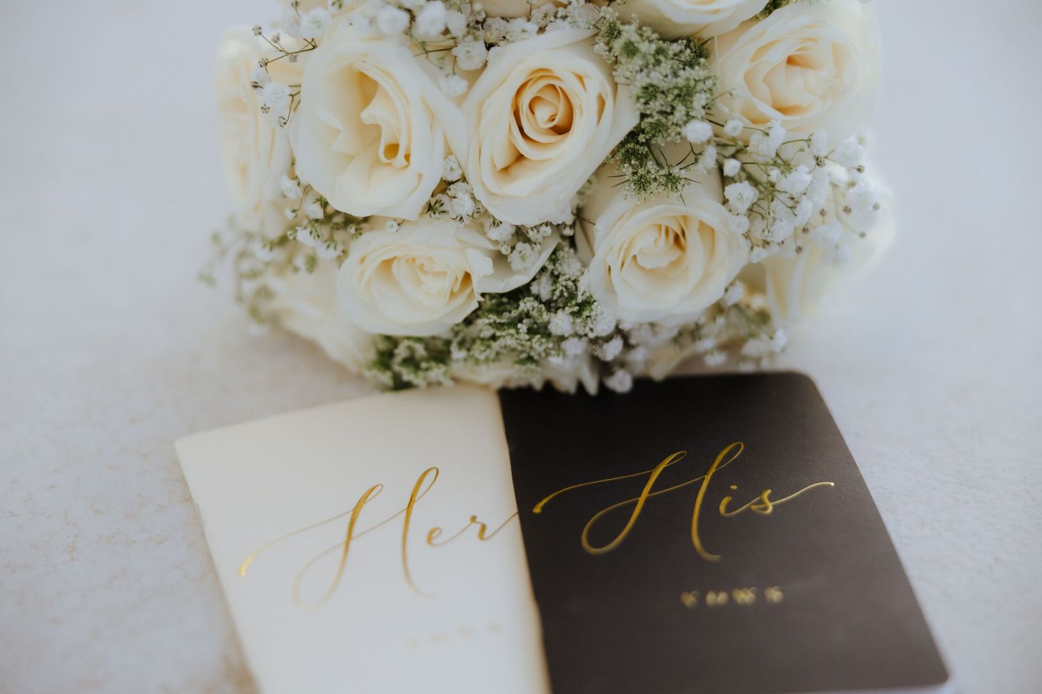 White rose bridal bouquet beside elegant gold script His and Hers wedding vow books.