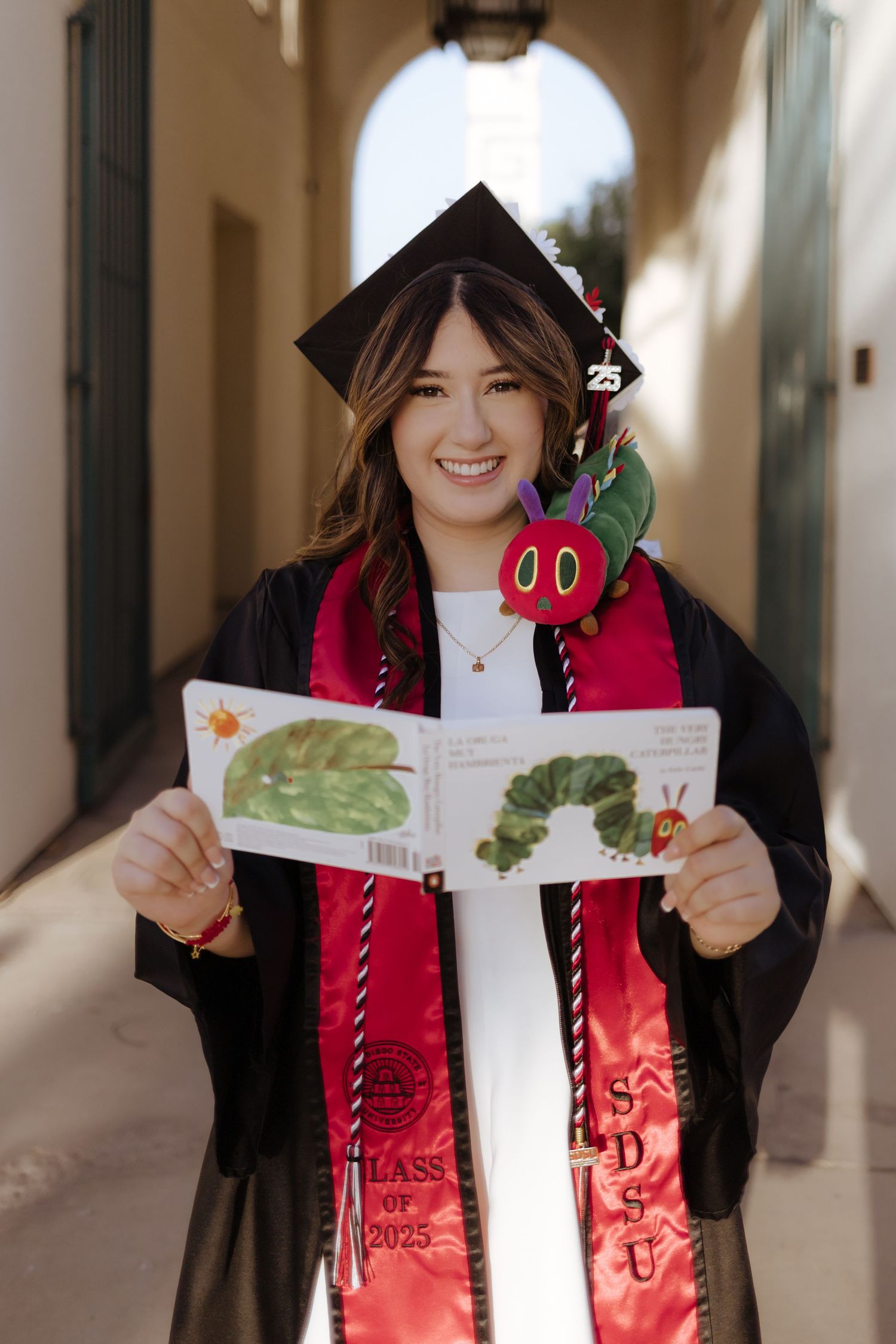 Smiling SDSU Class of 2025 graduate in cap and gown holds The Very Hungry Caterpillar book outdoors.