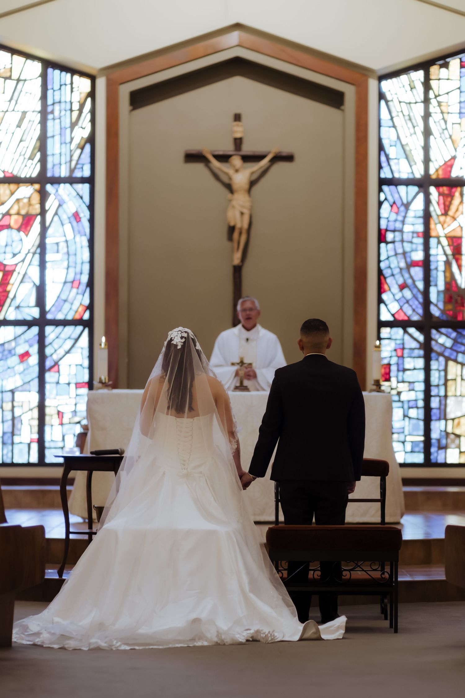 Bride and groom hold hands at the altar during a Catholic church wedding ceremony with stained glass windows.