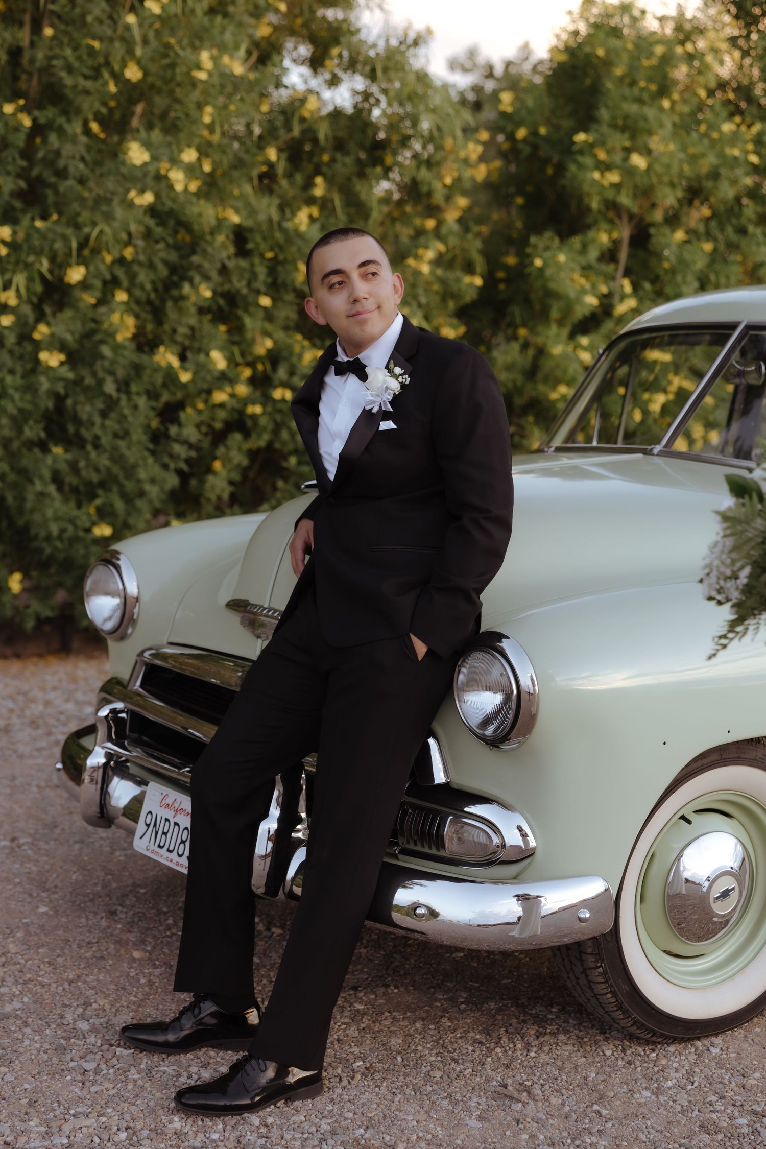 Dapper groom in black tuxedo with boutonniere leaning against a vintage mint green classic car.