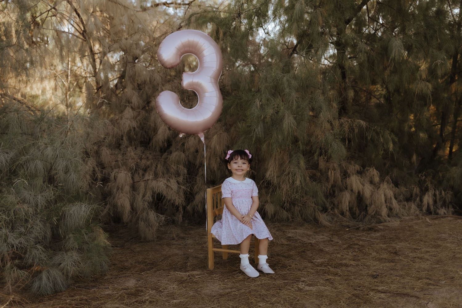 Young girl in white dress sitting on wooden chair holding pink number 3 balloon outdoors.